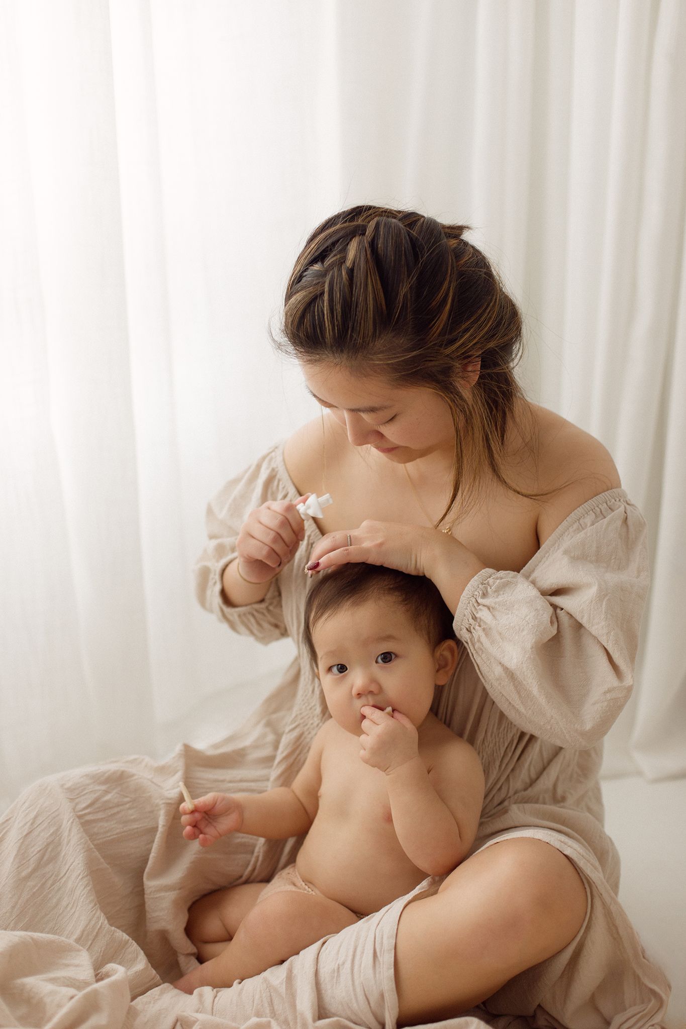 A mother in a flowing beige linen dress applies skincare drops to her baby's hair while the baby sits in her lap near sheer white curtains.