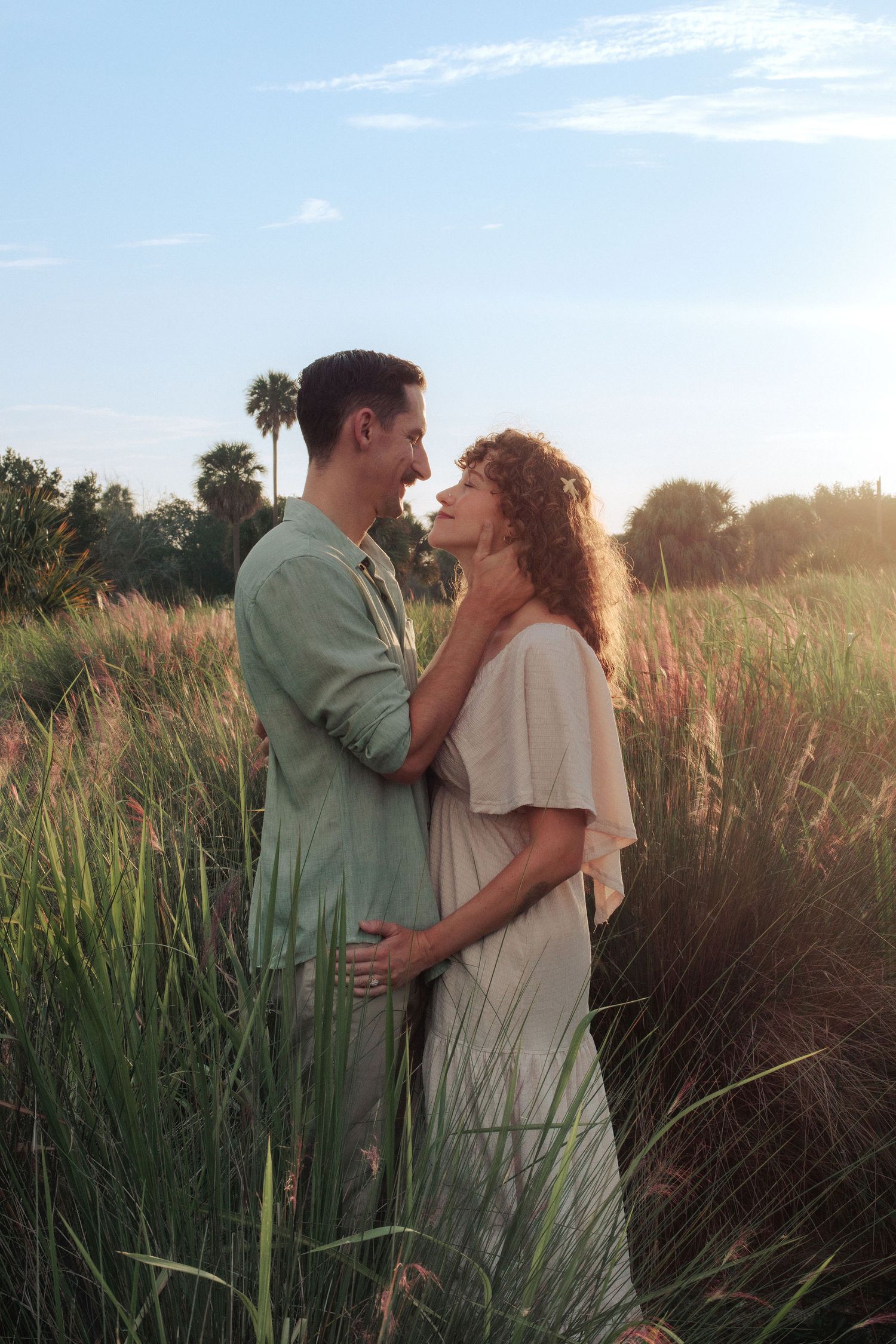 Couple embracing in a tall grass field at golden hour, with palm trees and a soft blue sky in the background.