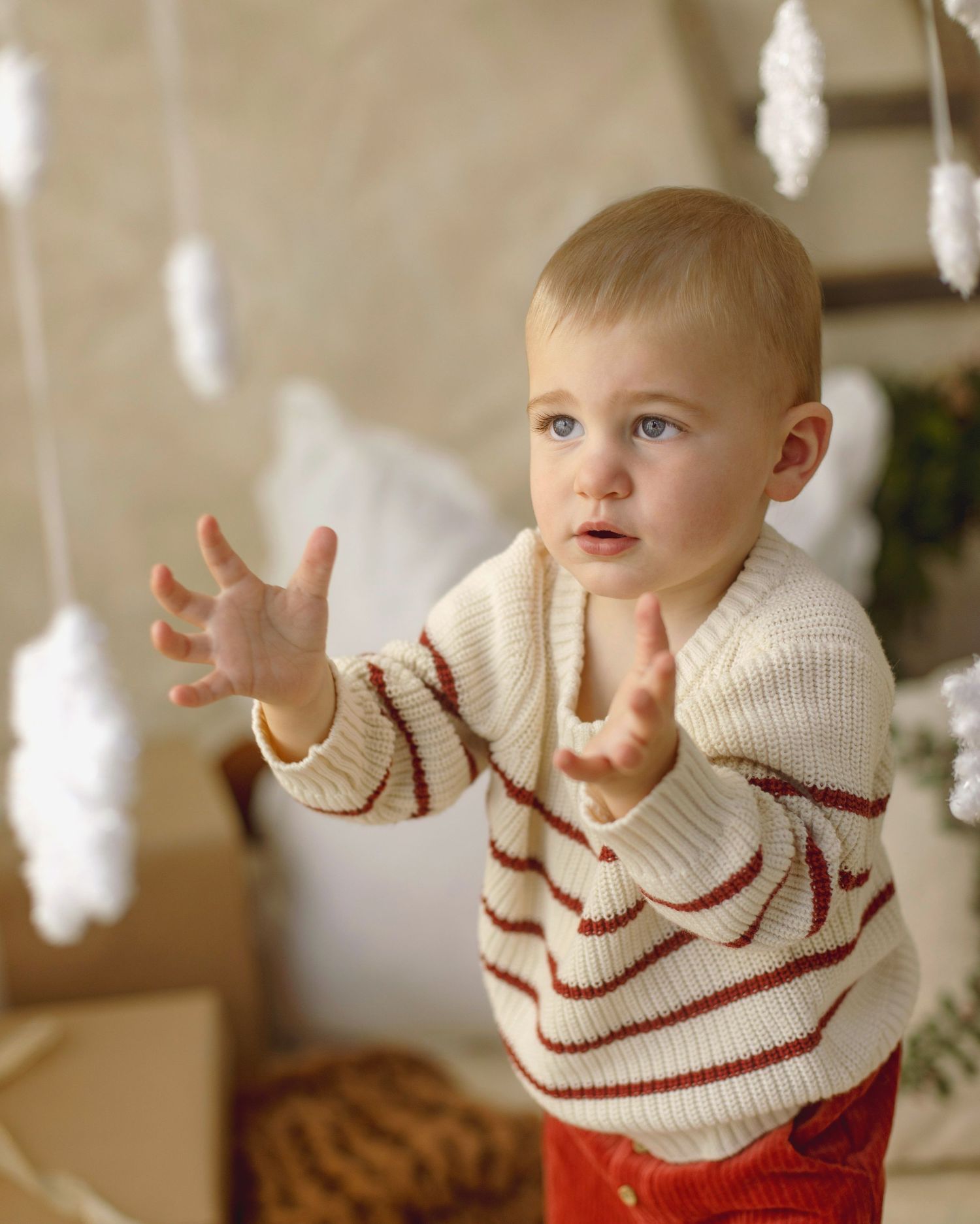 Adorable toddler in cream and red striped knit sweater reaching out hands toward hanging white decorations.
