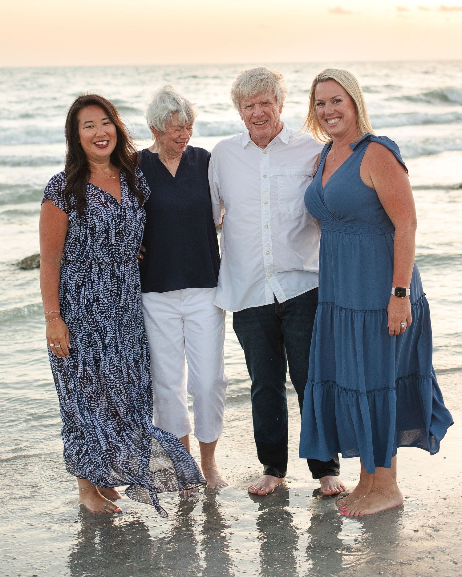 Four people smiling together on a beach at sunset, wearing blue and white outfits, waves in the background.