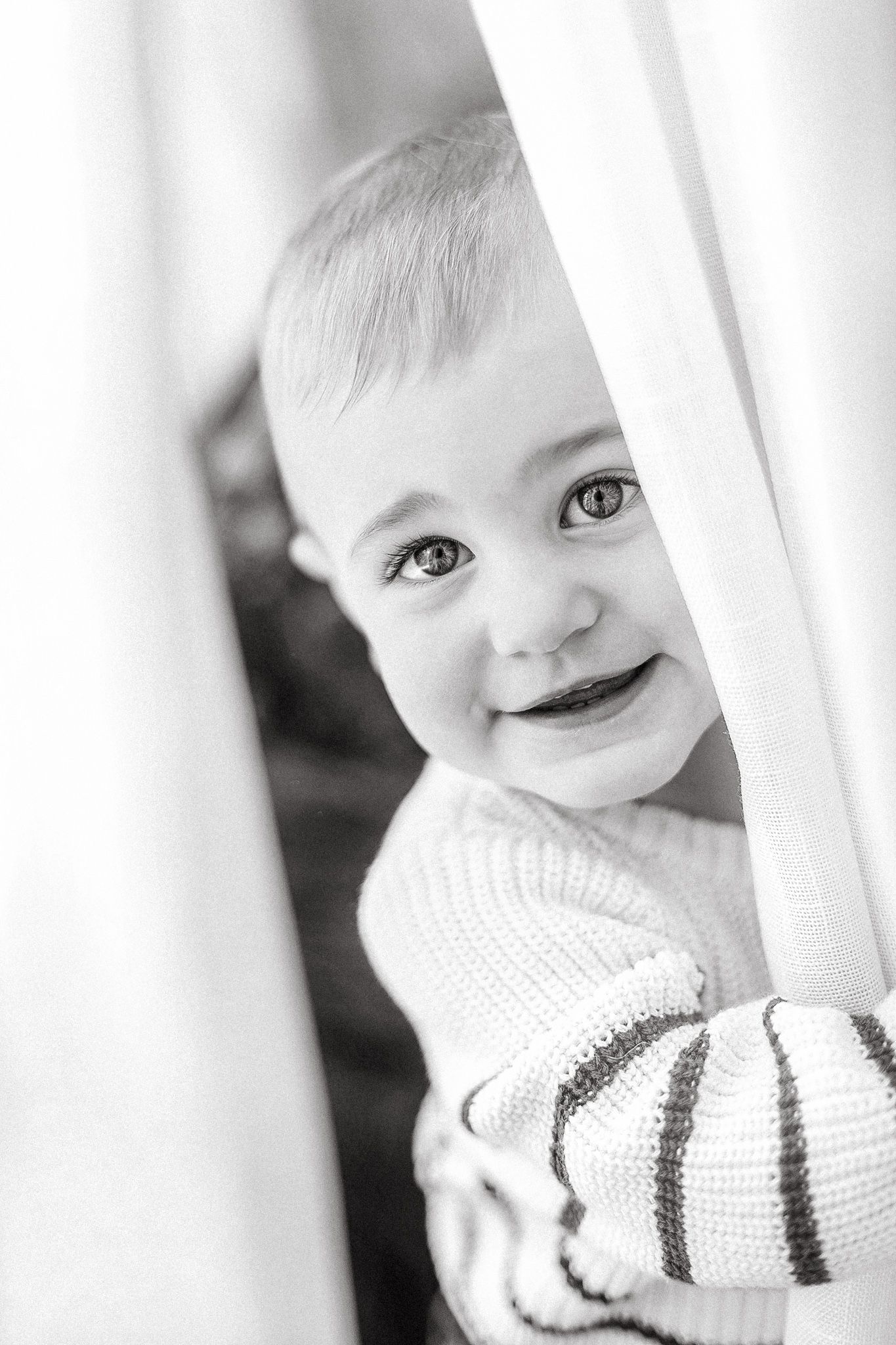 Black and white portrait of a smiling baby peeking through white curtains wearing a striped sweater.