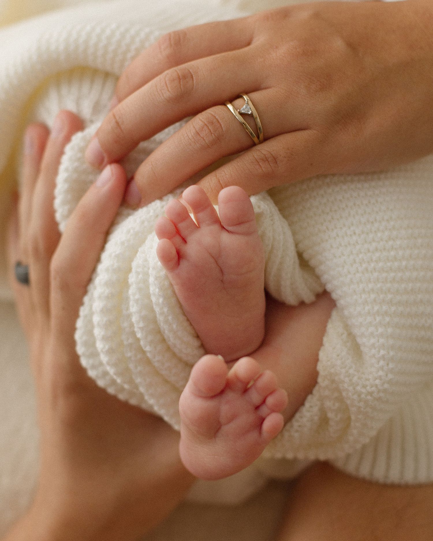 Close-up of newborn baby feet wrapped in white knit blanket held by mother wearing gold diamond ring.