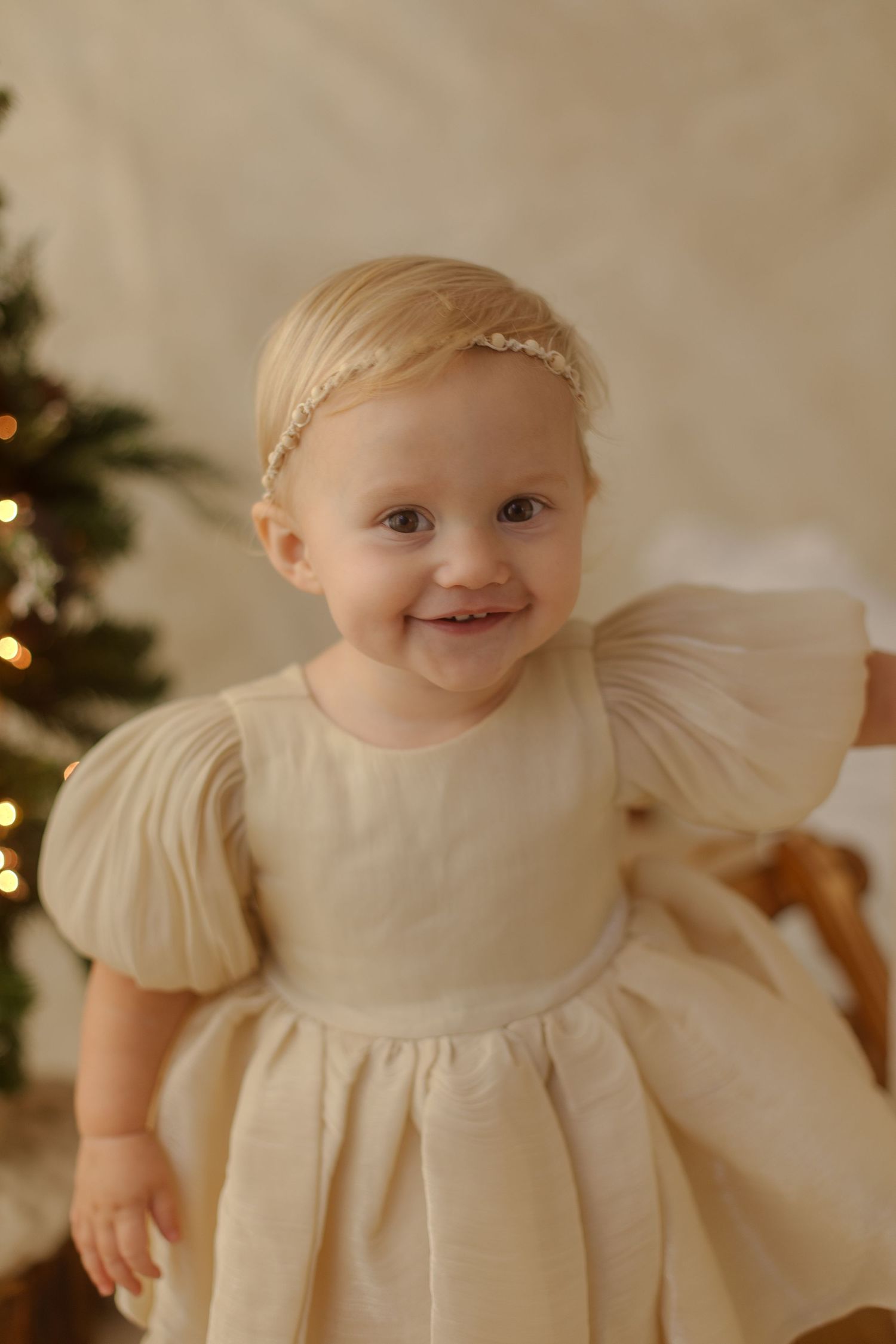 Smiling toddler girl in cream puff-sleeve dress with pearl headband near Christmas tree.