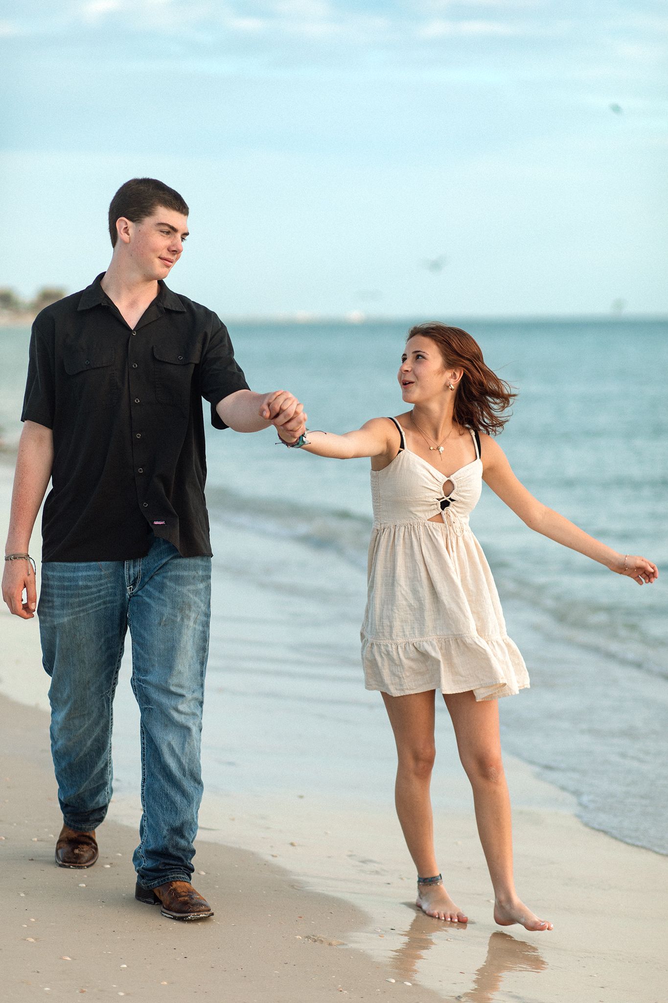 A young couple holds hands while walking along a sandy beach, with the woman in a white dress and man in a black shirt.
