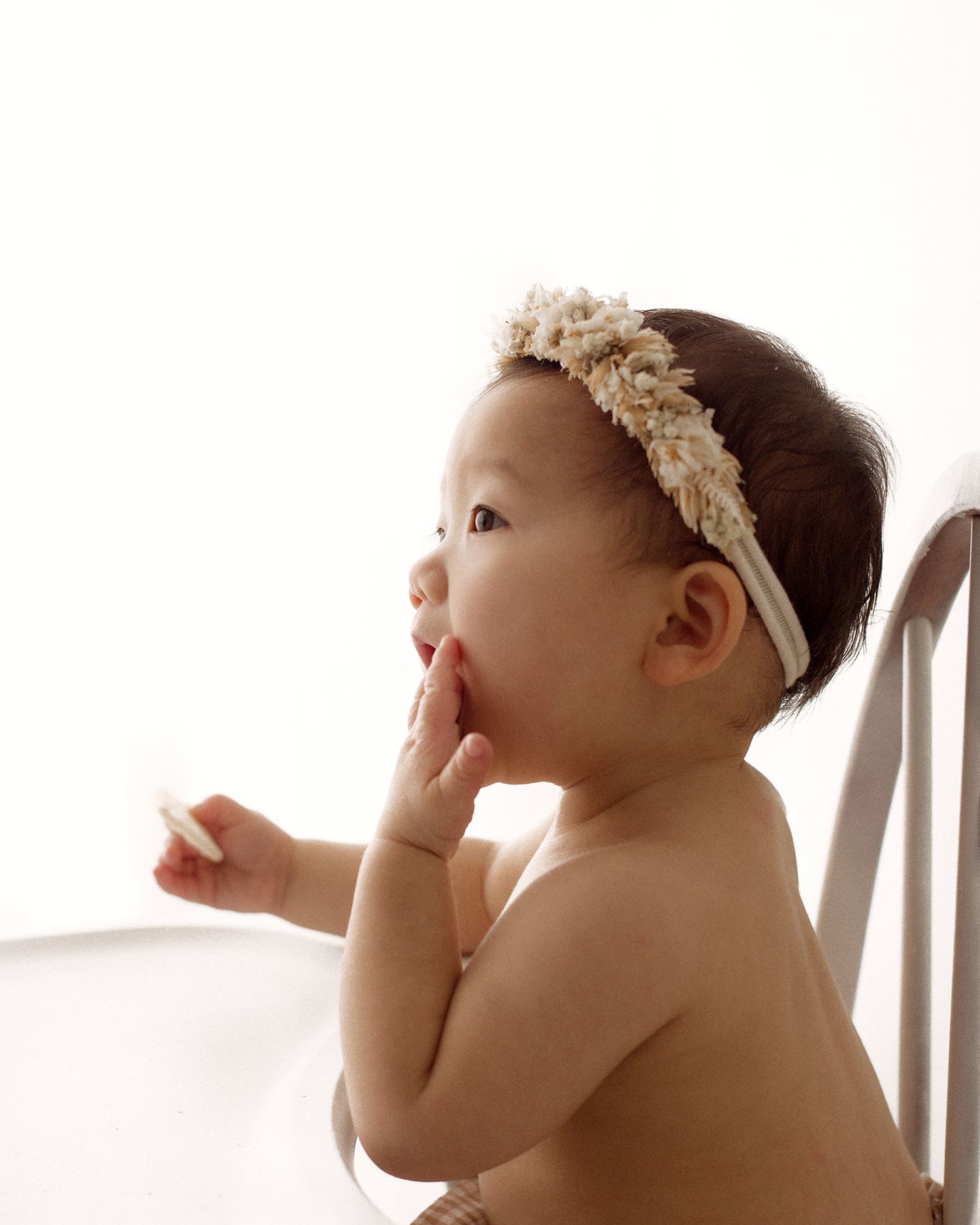 Baby in a floral headband sitting on a white chair, holding a small cookie and chewing on one hand against a bright background.