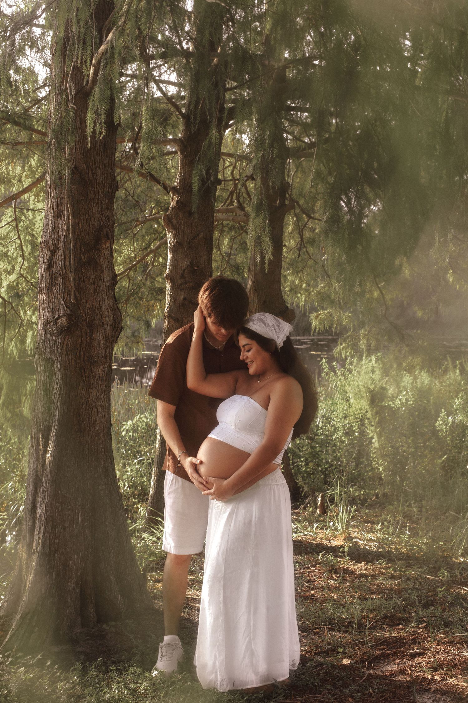 Loving couple standing under forest trees, pregnant woman in white dress gently cradling her growing baby bump.
