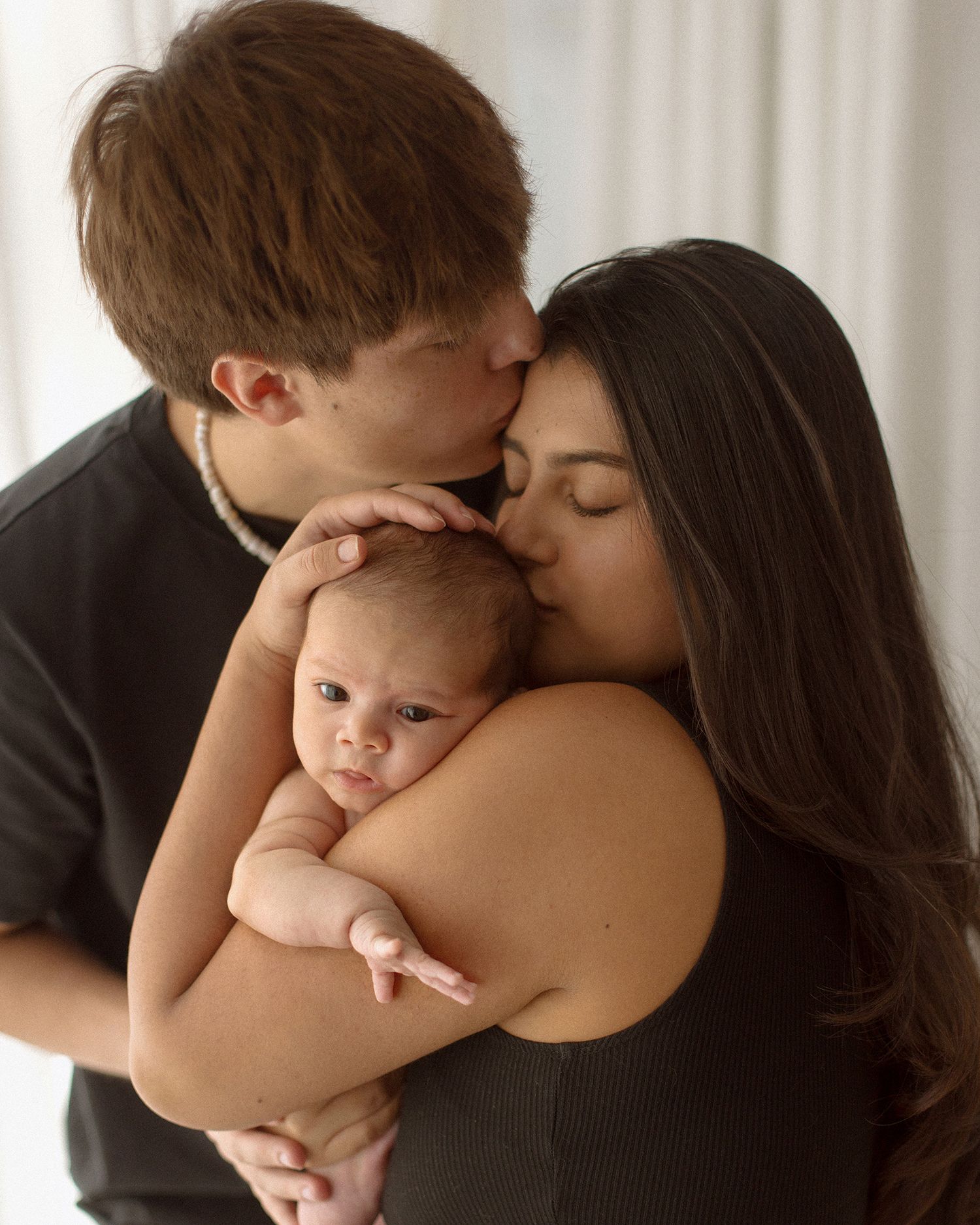 A young couple lovingly kisses their newborn baby, with the mother cradling the infant in a tender family portrait.