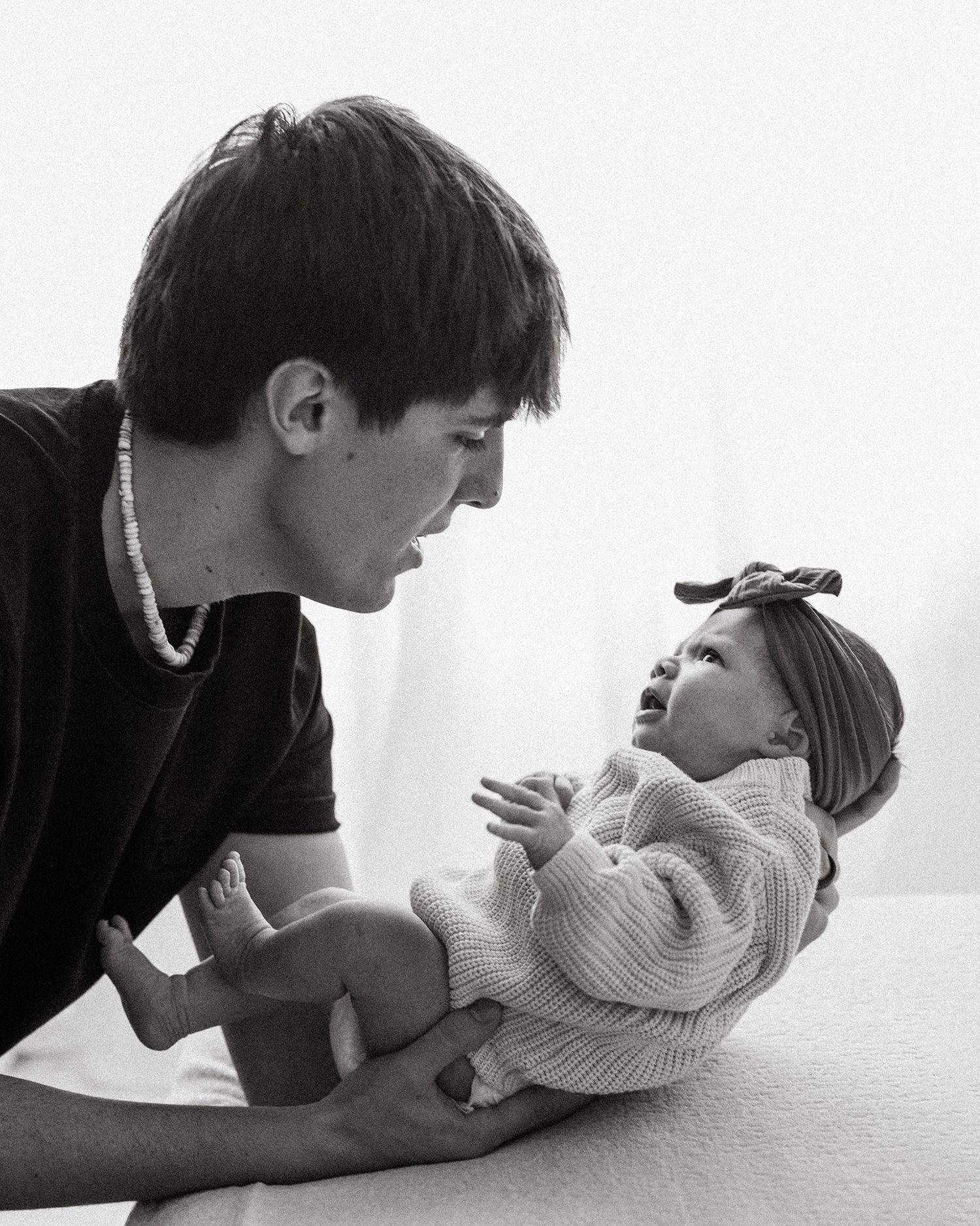 Black and white photo of a young man holding and smiling at a newborn baby wearing a knit sweater and bow headband.