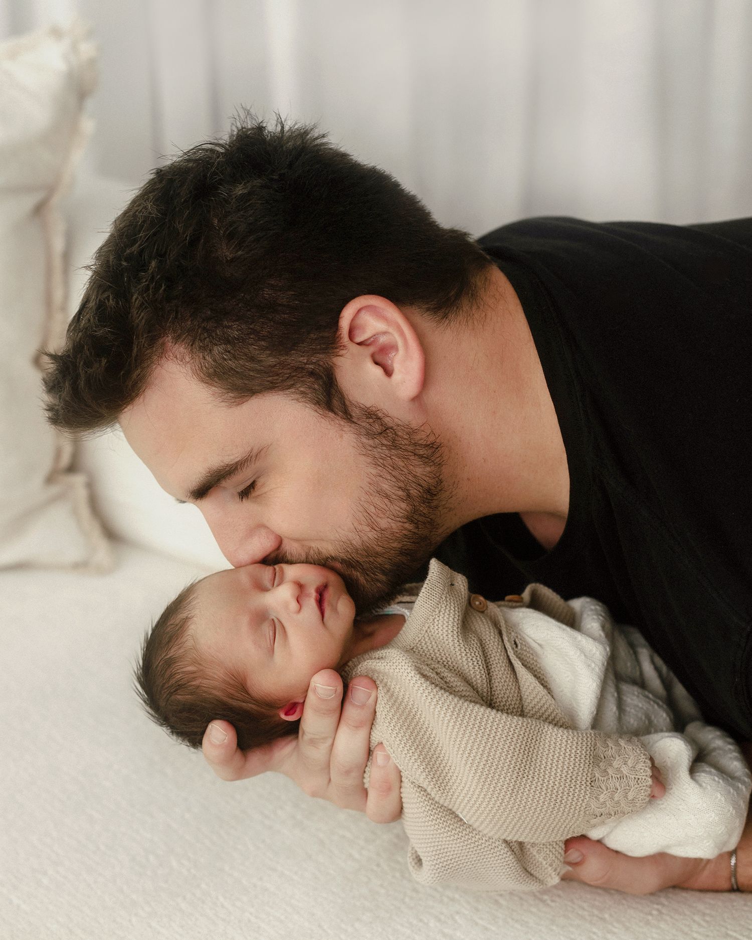 A bearded man in a black shirt tenderly kisses a sleeping newborn baby wrapped in a beige swaddle.