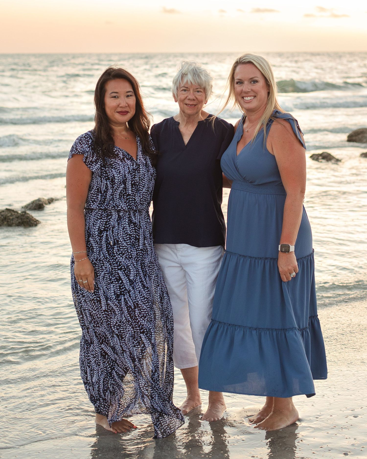 Three women smiling together on a beach at sunset, wearing blue outfits, standing barefoot in shallow water.