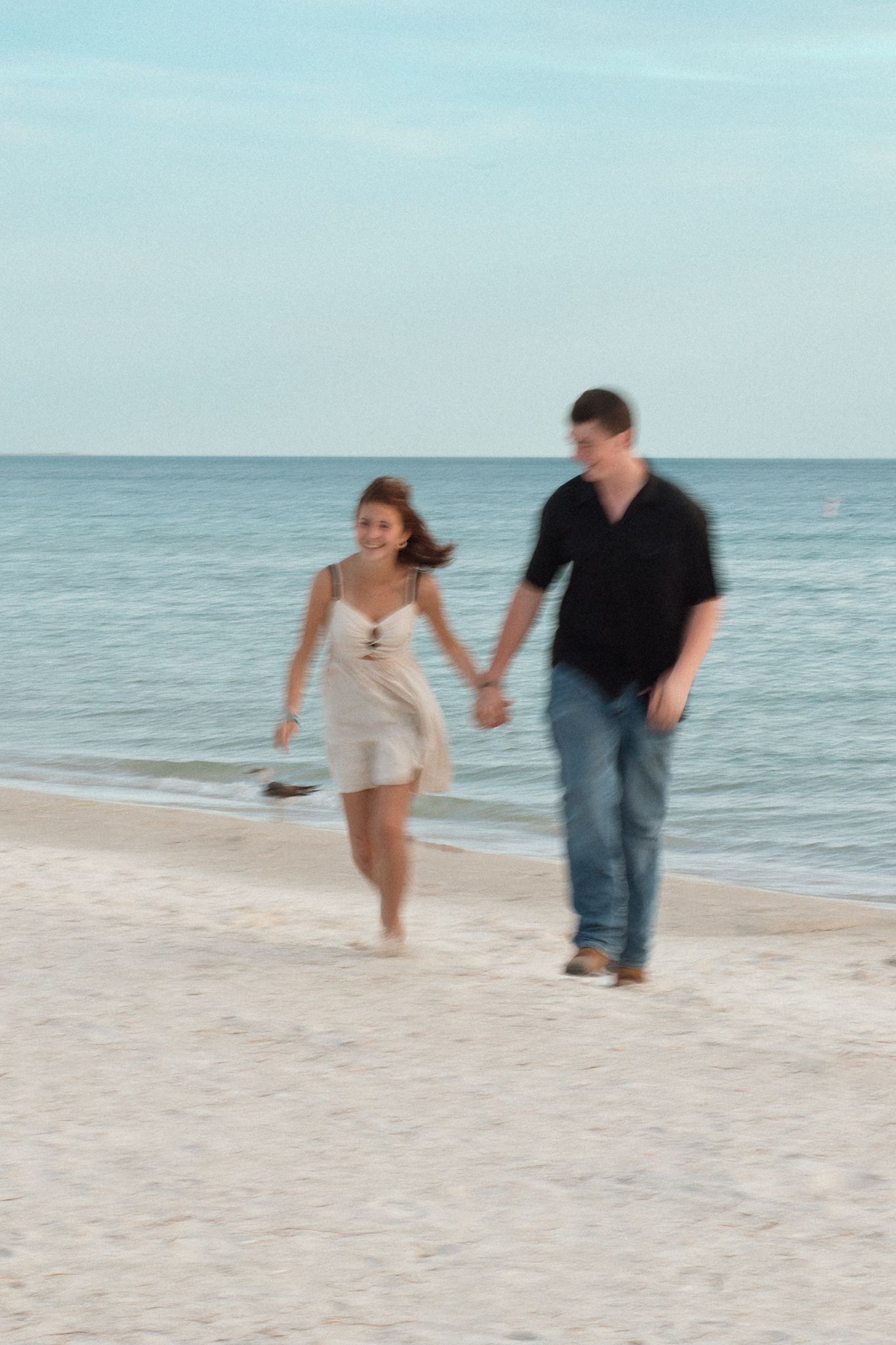 Couple holding hands walking on a sandy beach near the ocean shoreline on a clear day.