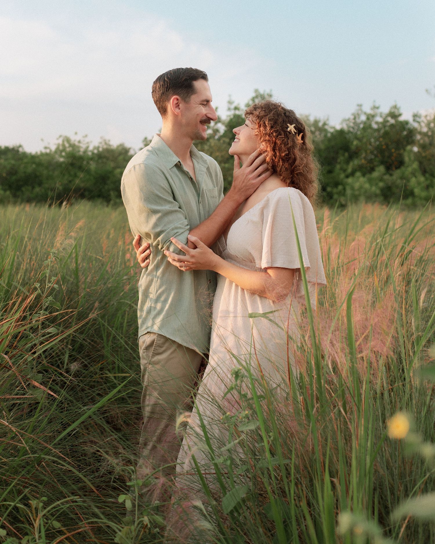 Smiling couple embracing in a tall grass field at golden hour, the woman in a flowing dress and the man in a green shirt.