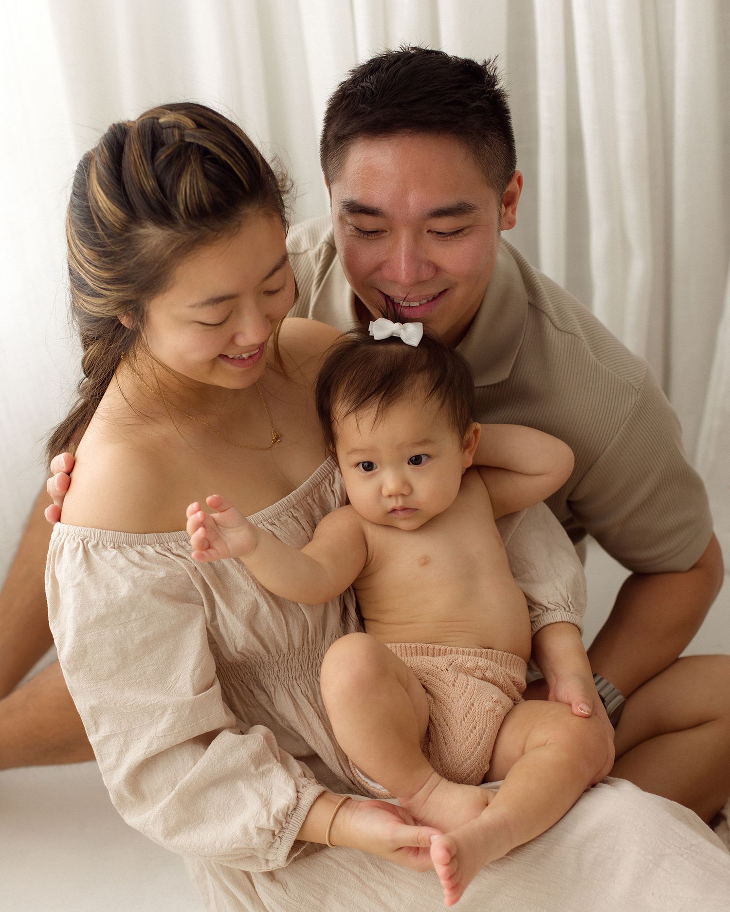 A smiling woman and man in neutral tones gently hold a baby wearing a white hair bow in a bright, soft family portrait.