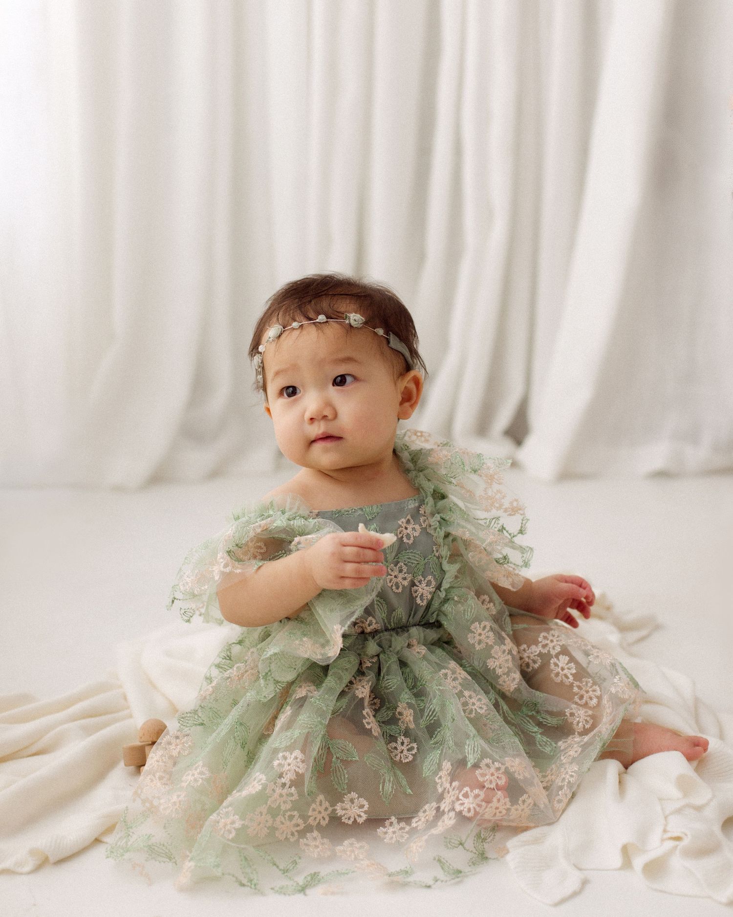 Baby girl in a green tulle dress with embroidered flowers and a floral headband sits on a white blanket against soft white drapes.
