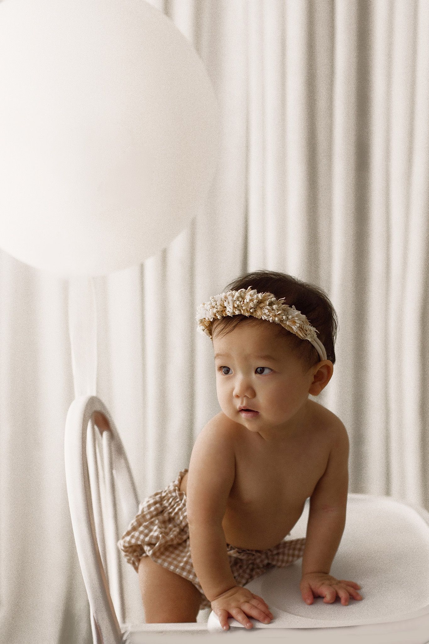 Baby in a gingham romper and floral headband crawls on a white chair beside a large white balloon against soft cream curtains.