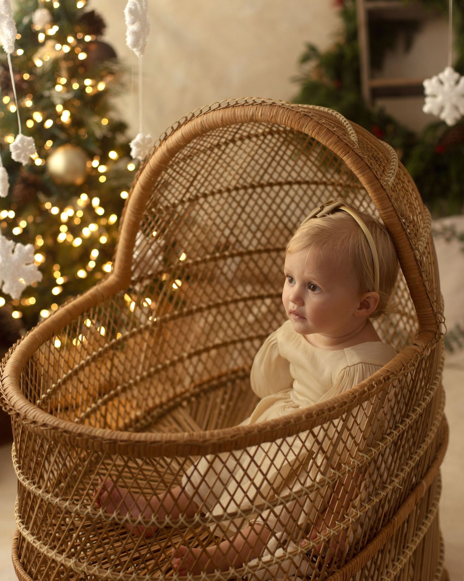 Baby in a wicker Moses basket beside a decorated Christmas tree with warm glowing lights.