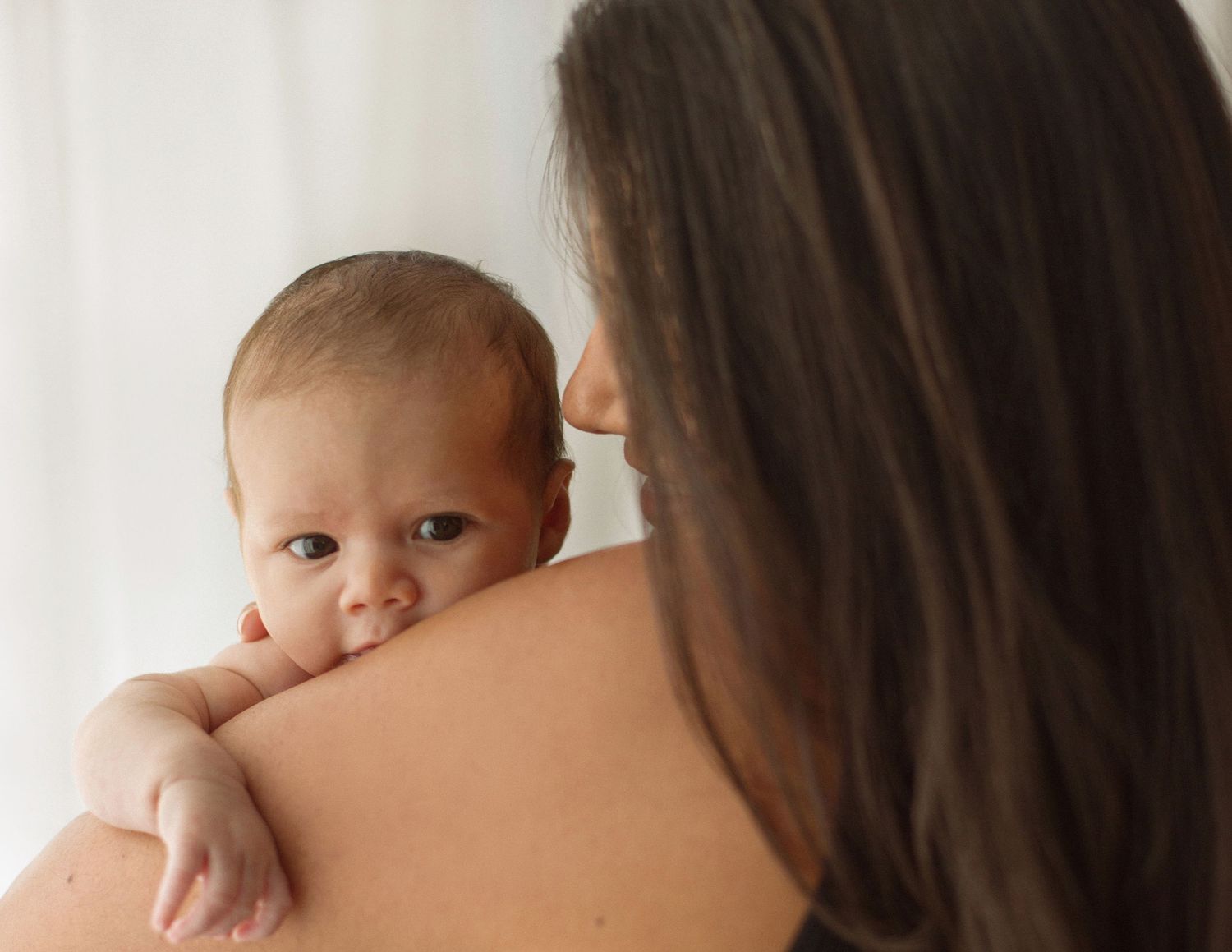 Mother holding newborn baby on her shoulder, infant looking over mom's back in soft natural light.