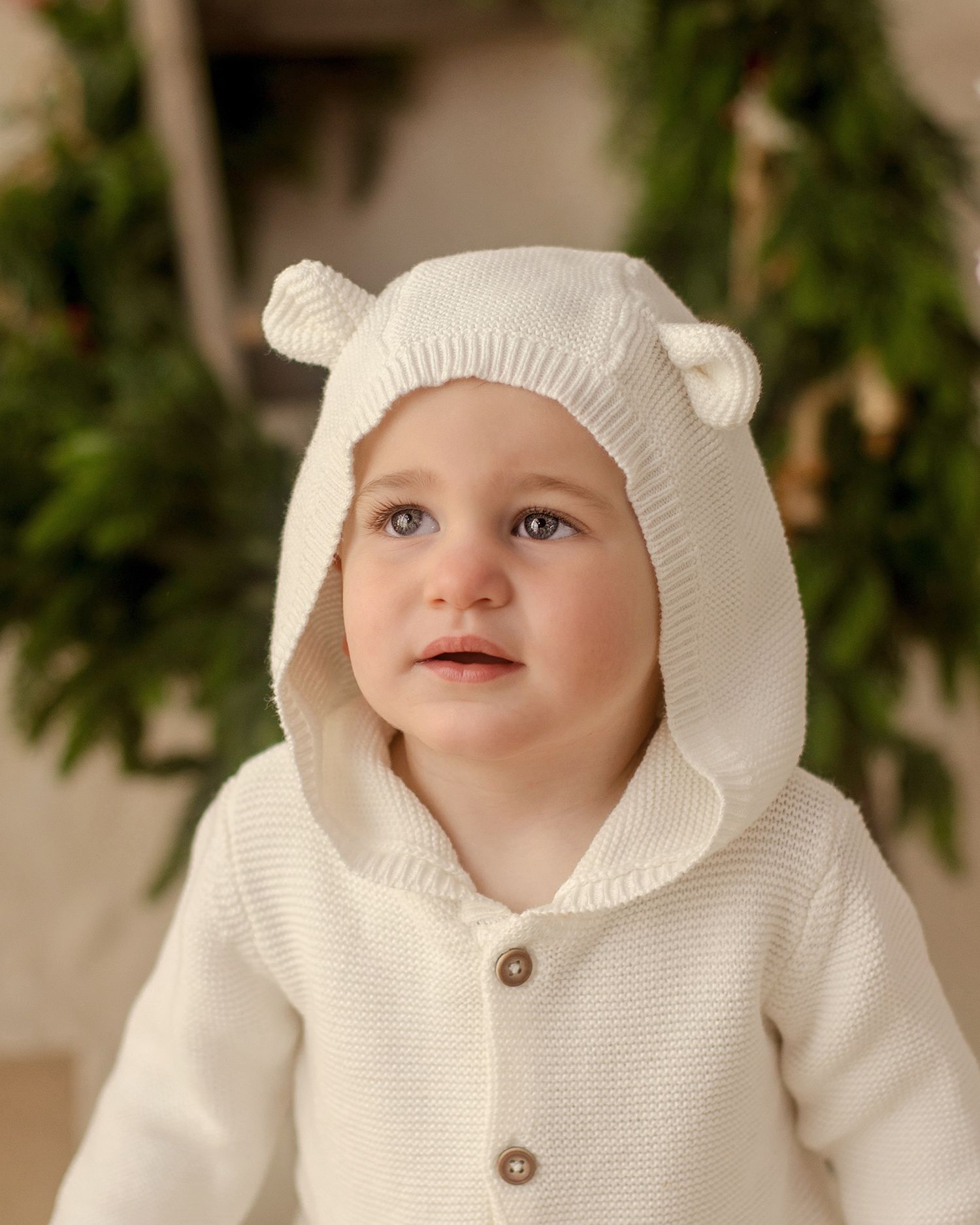 Baby in white knit bear ear hooded sweater with wood buttons, sitting in front of green holiday garland.