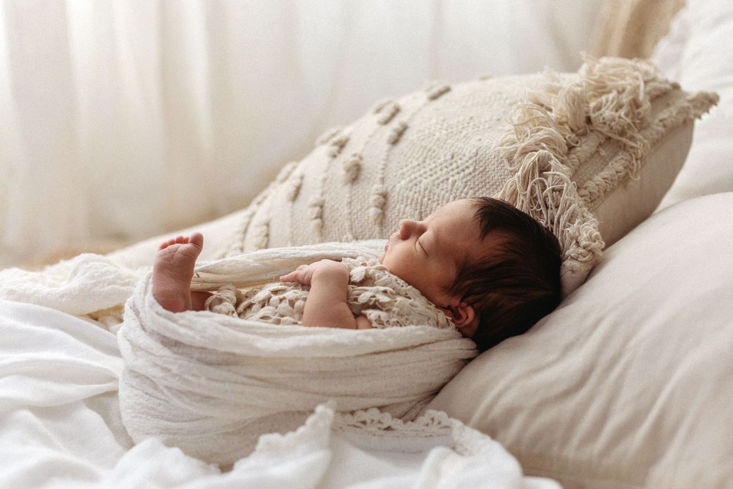 Newborn baby sleeping peacefully wrapped in white lace swaddle on soft bedding with cream boho pillow.