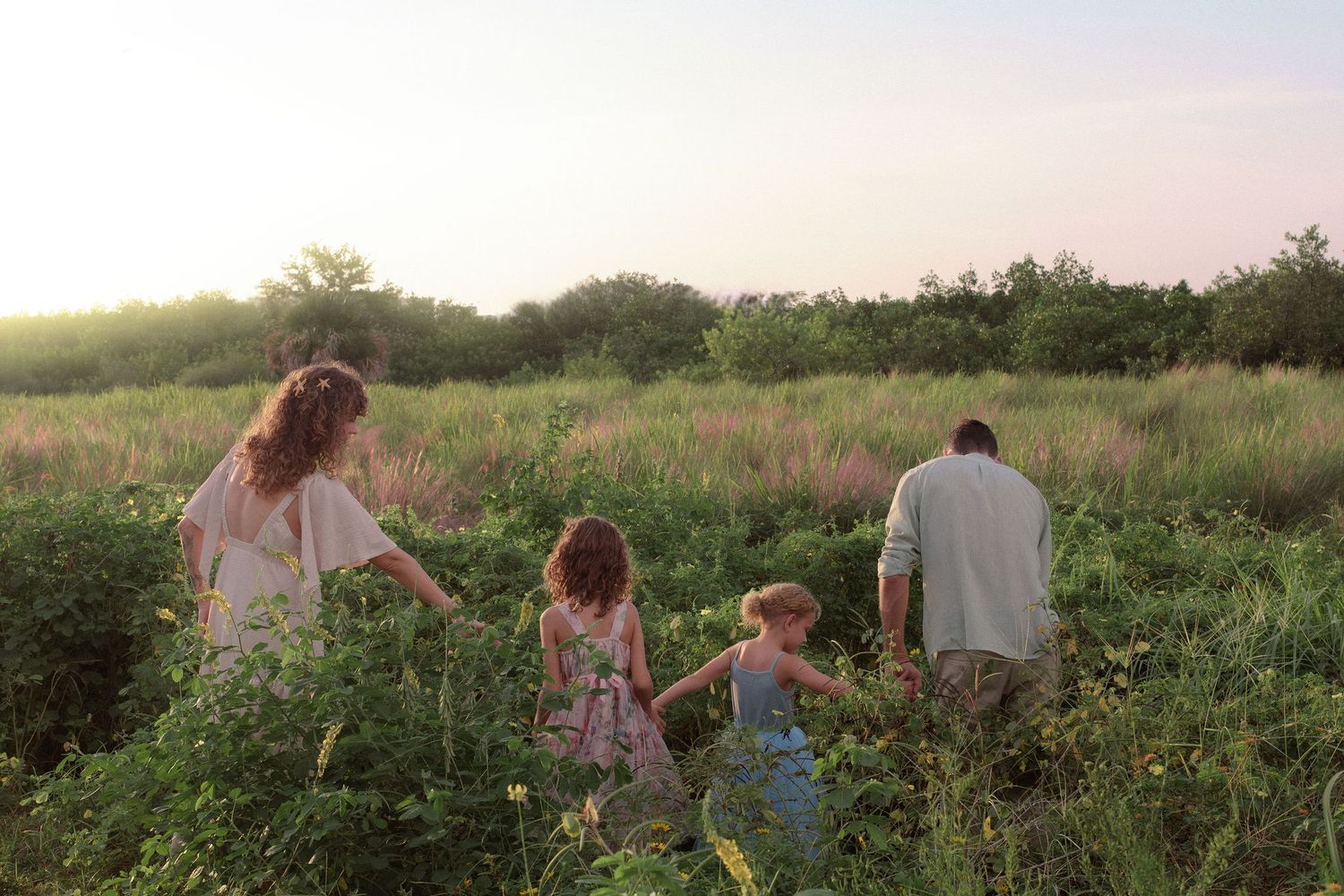 Family of four with two young girls walking through a lush green meadow of wildflowers and tall grass at golden sunset.