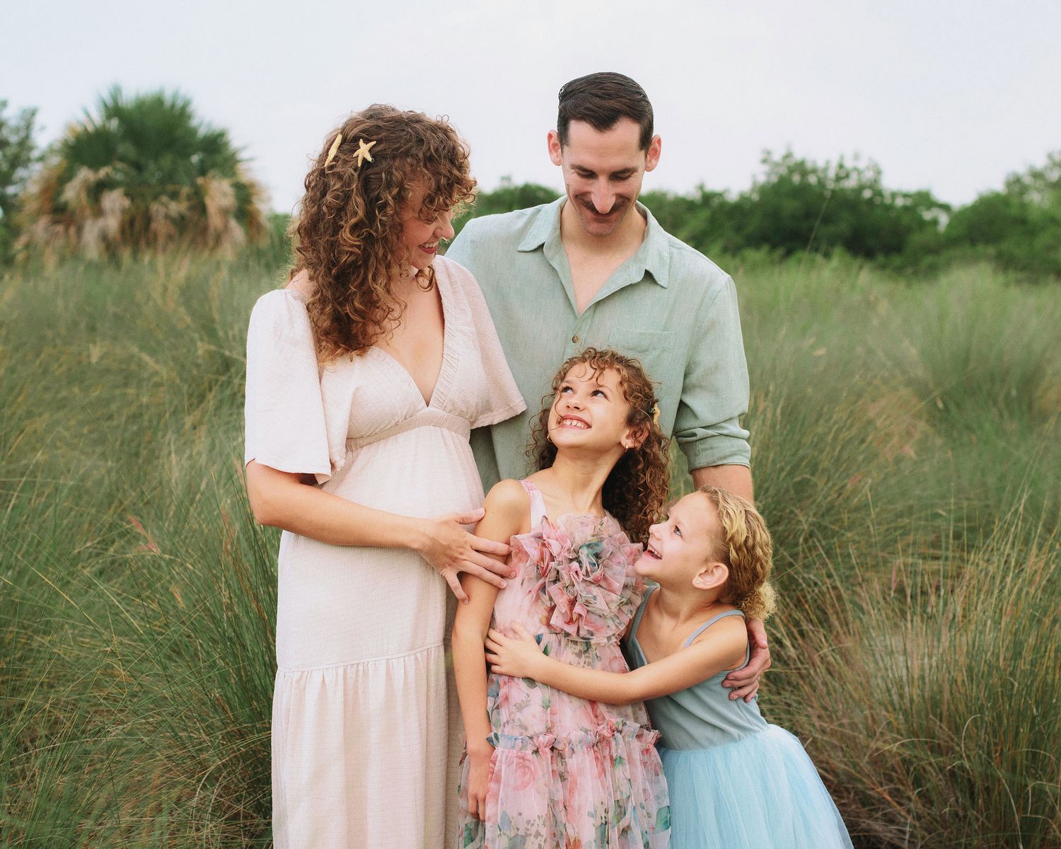 Smiling family of four embracing in tall green grass during an outdoor portrait session, with two young girls in pastel dresses.