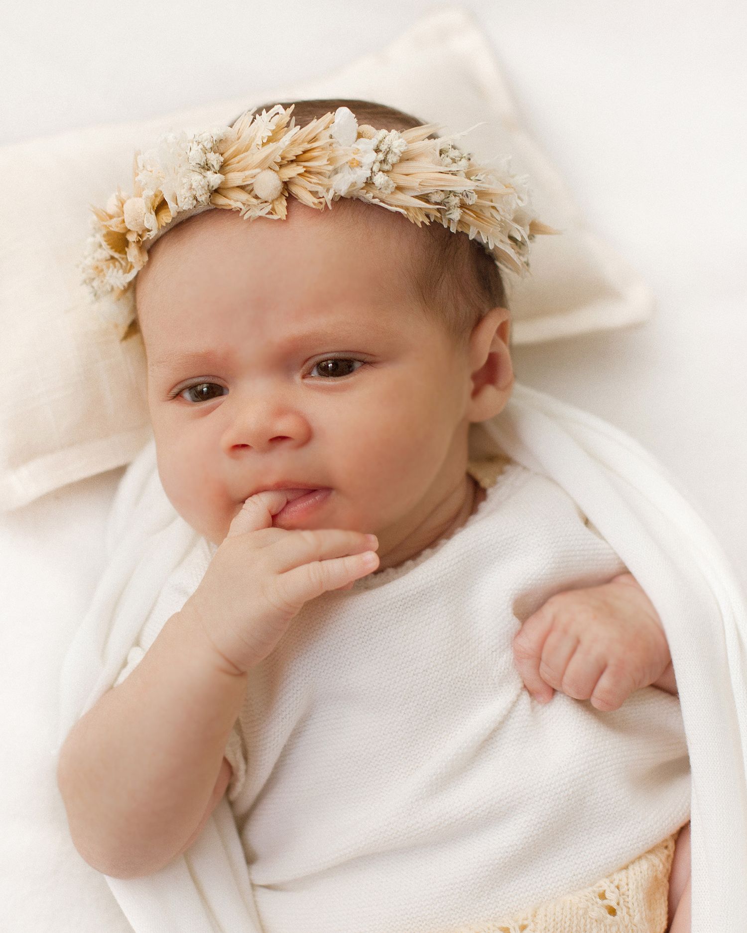 A newborn baby wearing a delicate floral crown lies on a white pillow, with fingers near mouth.
