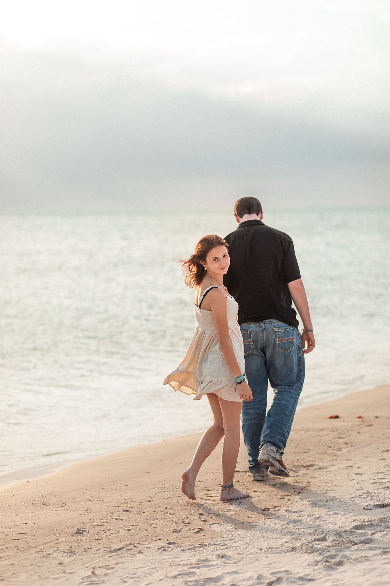 Couple walking on a sandy beach at sunset, woman in white dress looks back smiling, man in black shirt and jeans.