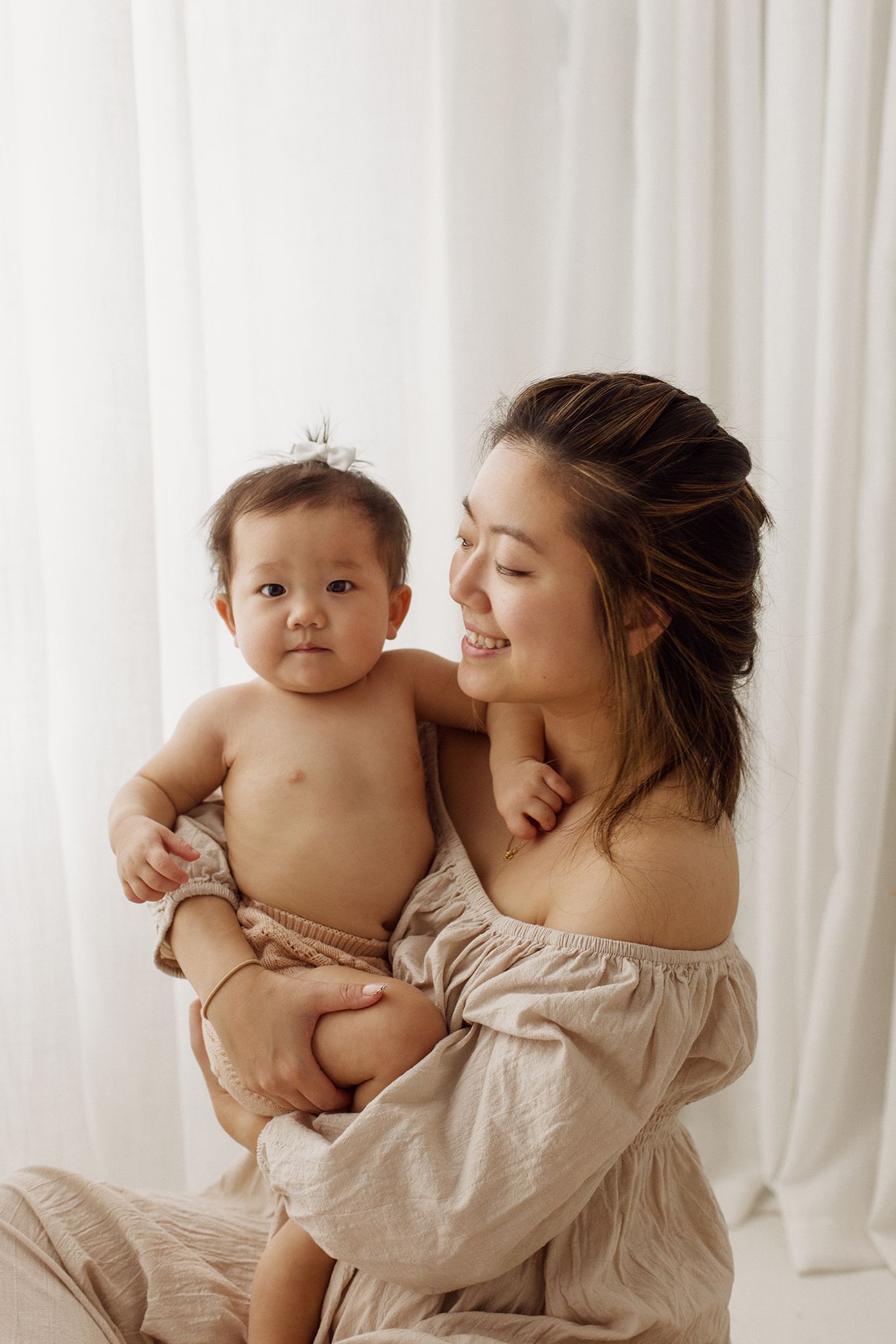 Smiling woman in an off-shoulder neutral linen outfit holds a baby with a small hair bow against soft white curtains in a bright studio.
