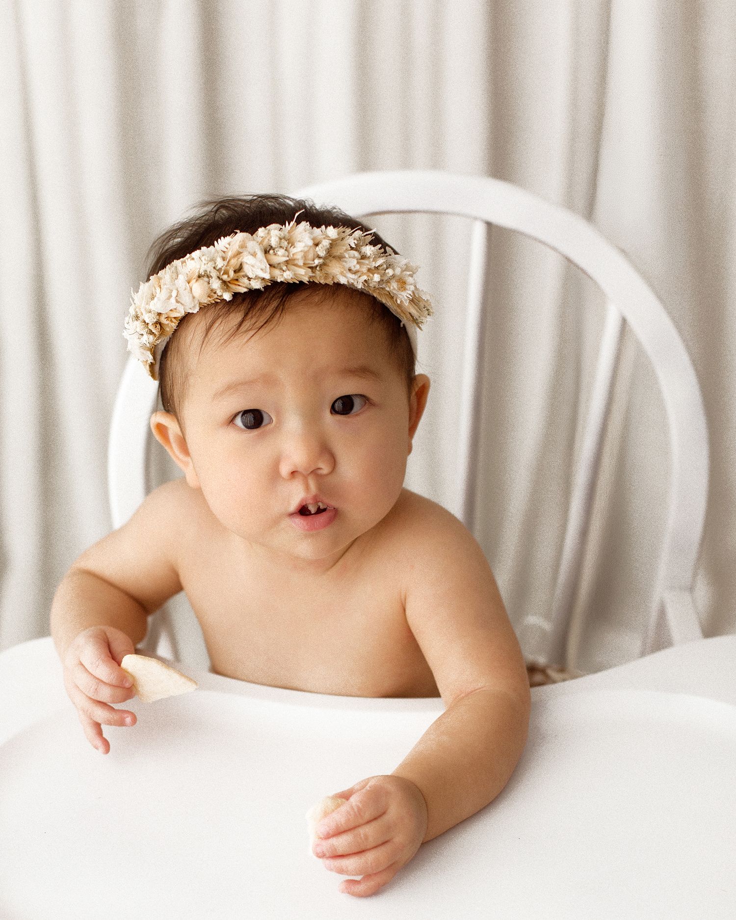 Baby wearing a cream floral headband sits in a white high chair, holding small snacks against a soft white curtain backdrop.