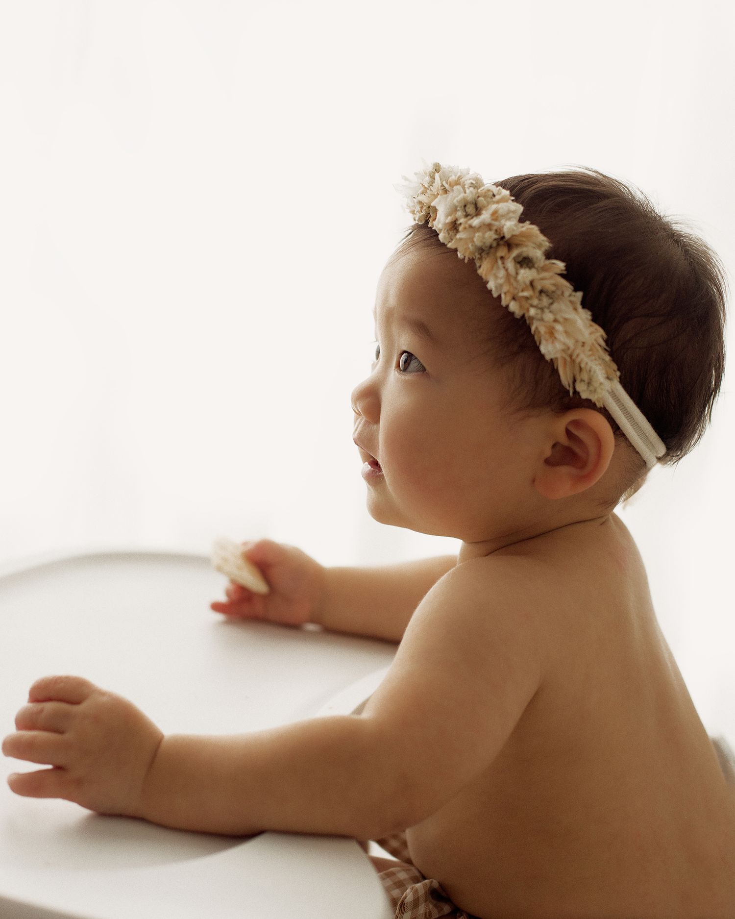 Baby wearing a floral headband sits at a white high chair tray, holding a small snack and looking to the side in soft natural light.