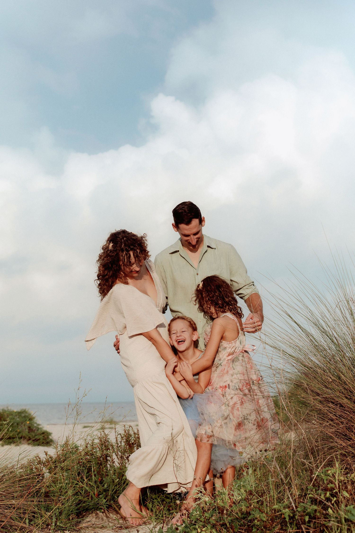 A joyful family of four laughs and embraces among beach dune grass, with the ocean and a cloudy sky behind them.