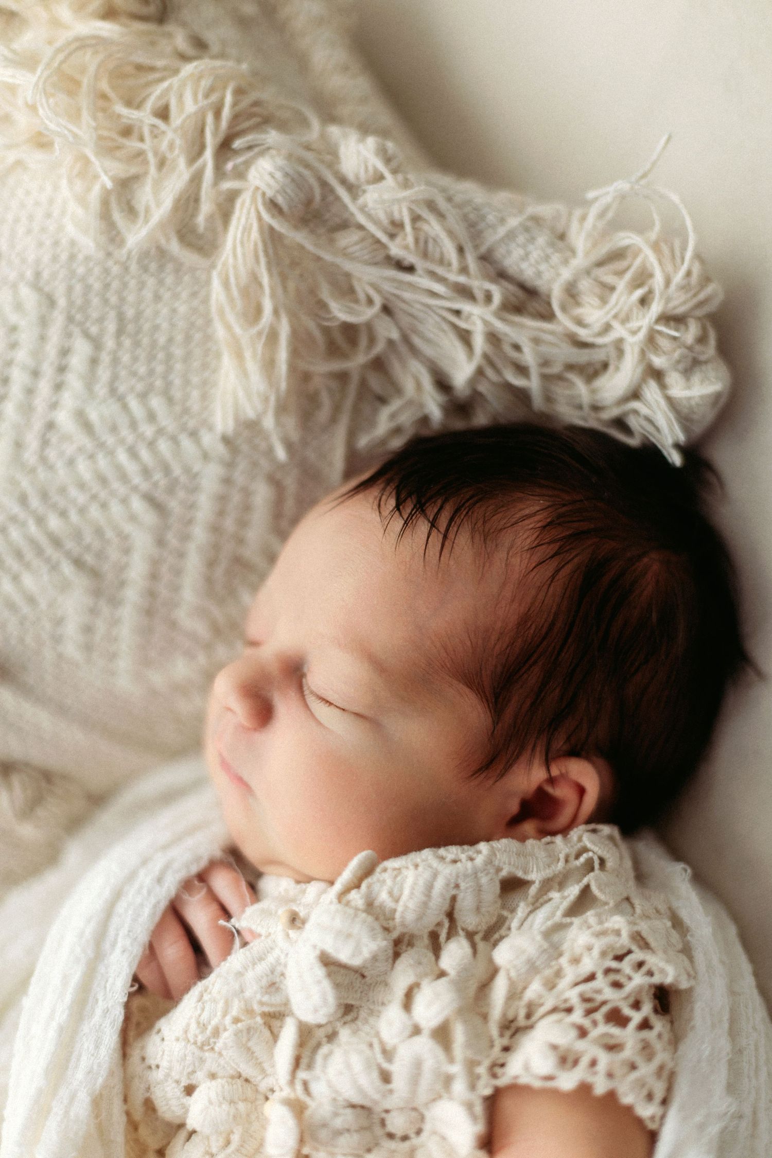 Sleeping newborn baby with dark hair wrapped in white lace blanket on soft cream textured background.