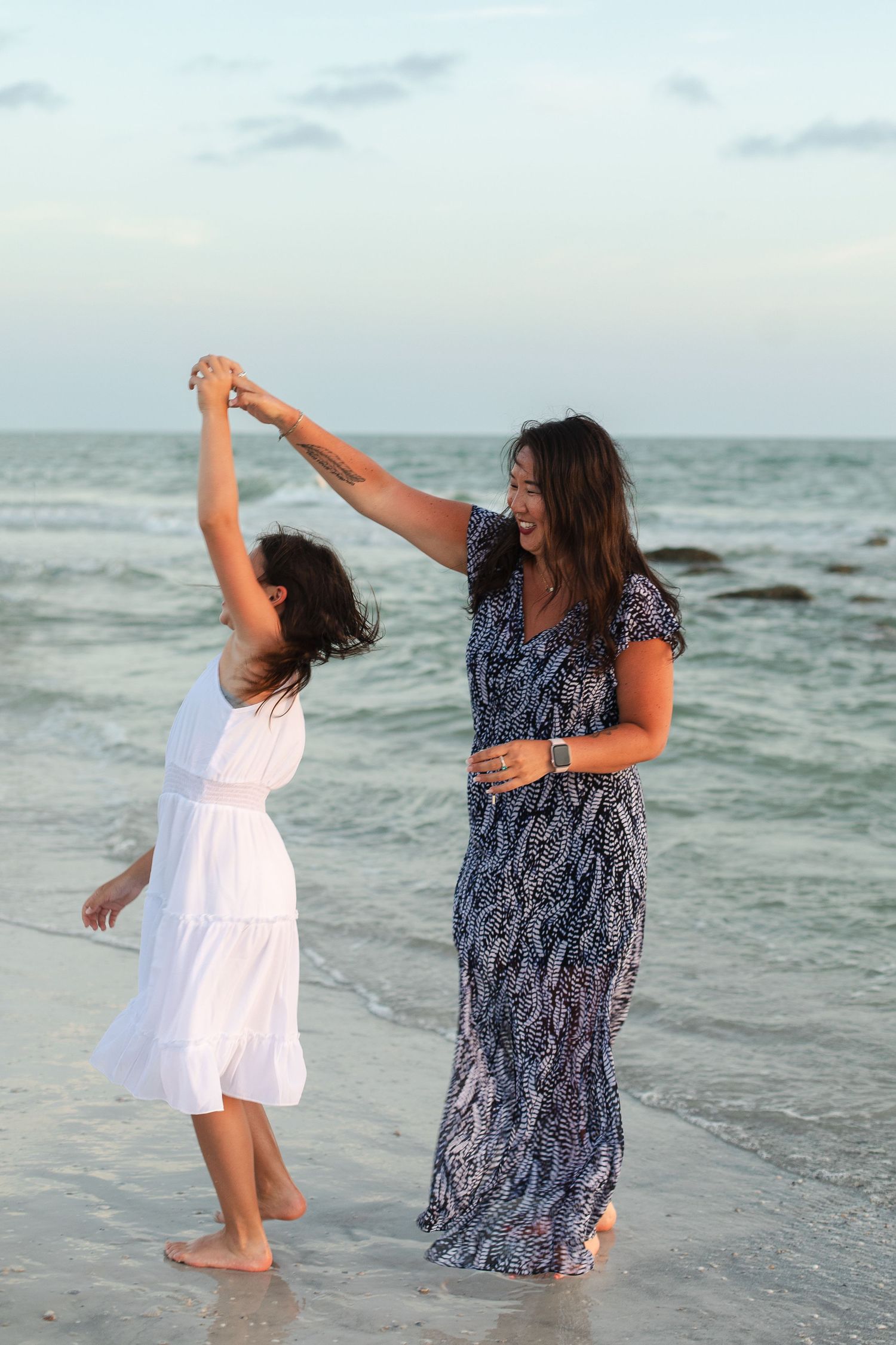 A woman and young girl dance joyfully on a beach at sunset, holding hands with waves in the background.