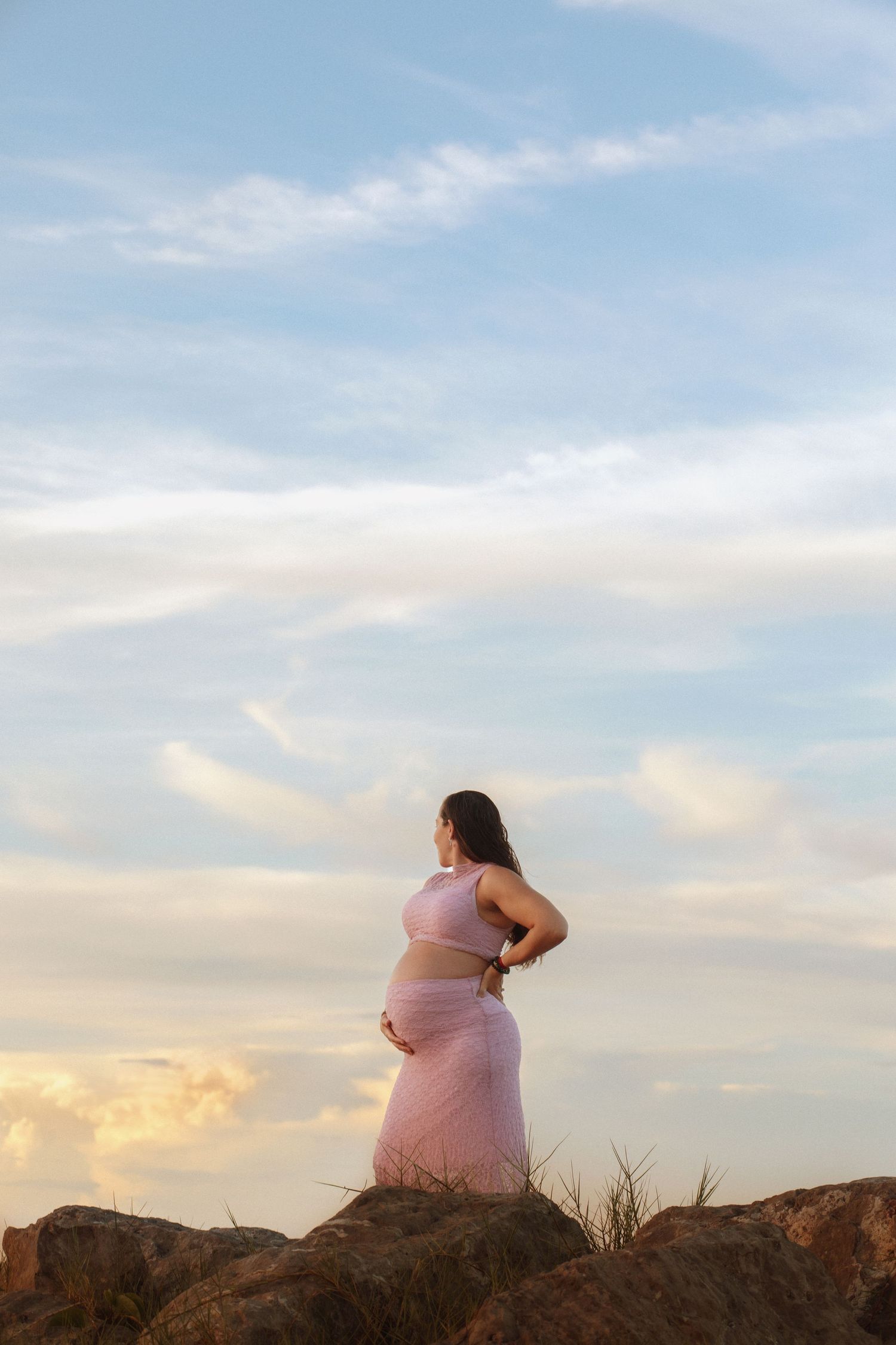 Pregnant woman in pink outfit standing on rocks against a serene sunset sky, cradling her baby bump.
