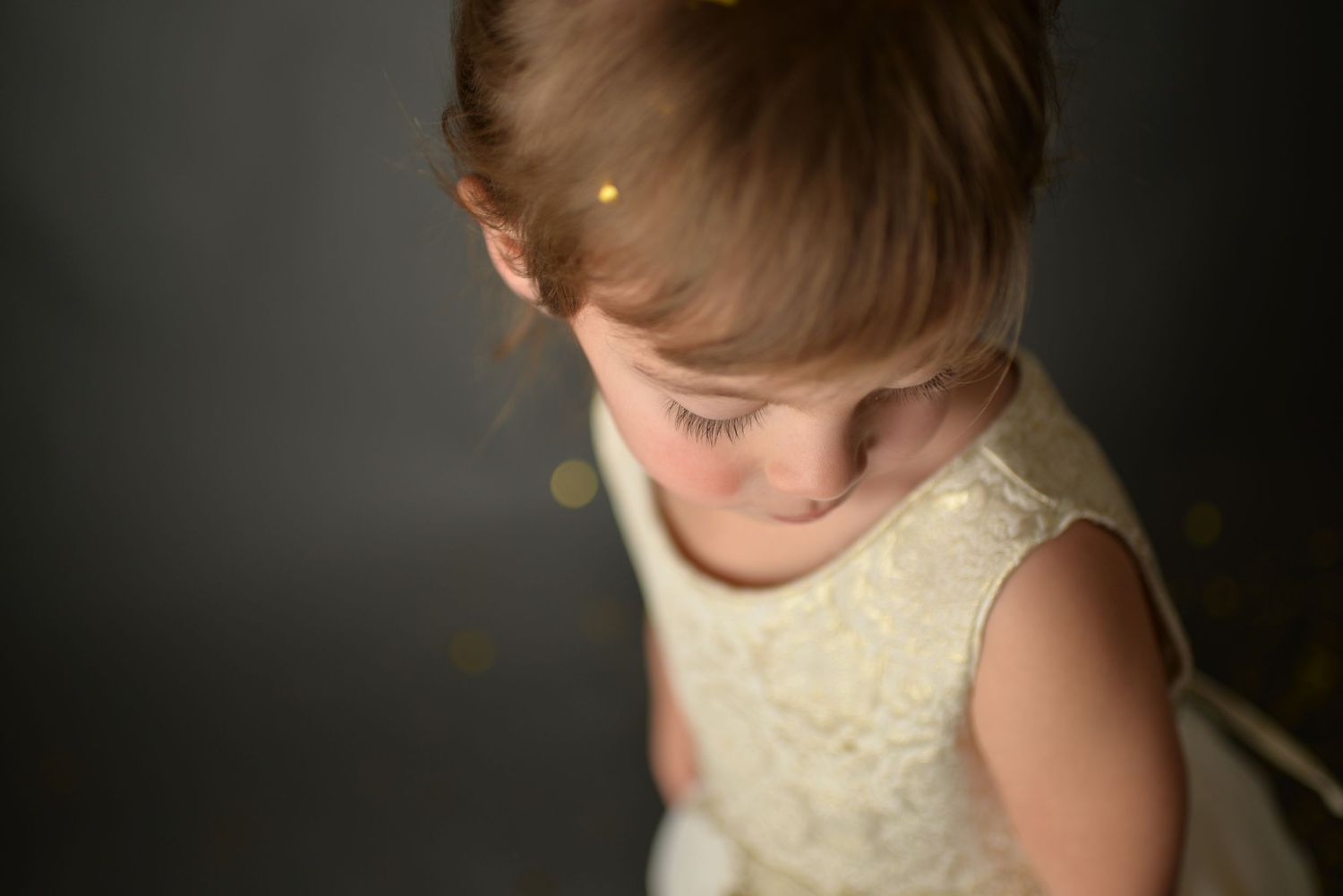 Young girl in white lace dress looking down, soft bokeh lights in dark background.