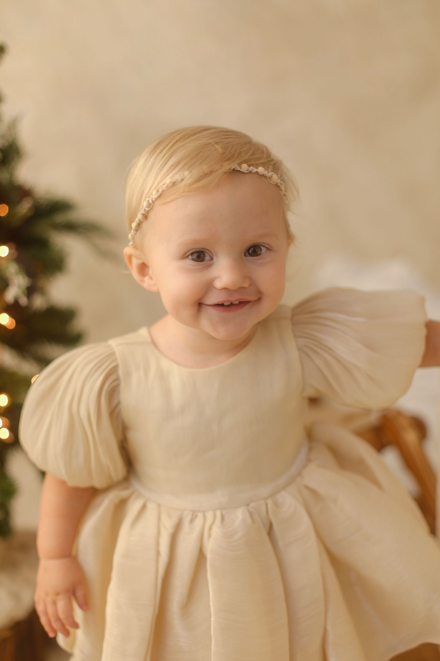 Smiling baby girl in cream puff-sleeve dress with pearl headband poses near a glowing Christmas tree.
