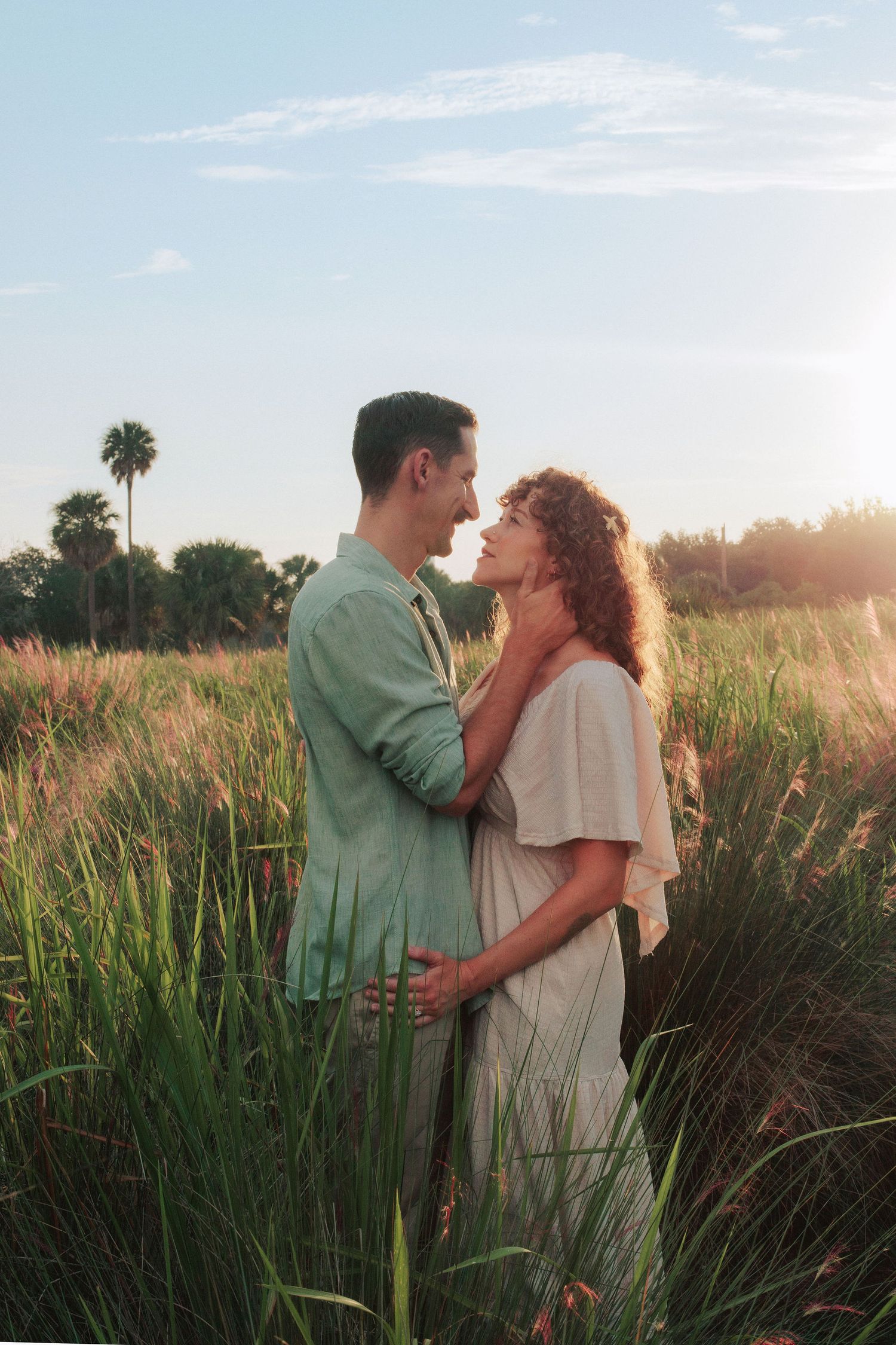 A couple embraces in a tall grass field at golden hour, with palm trees and a soft sunset sky behind them.