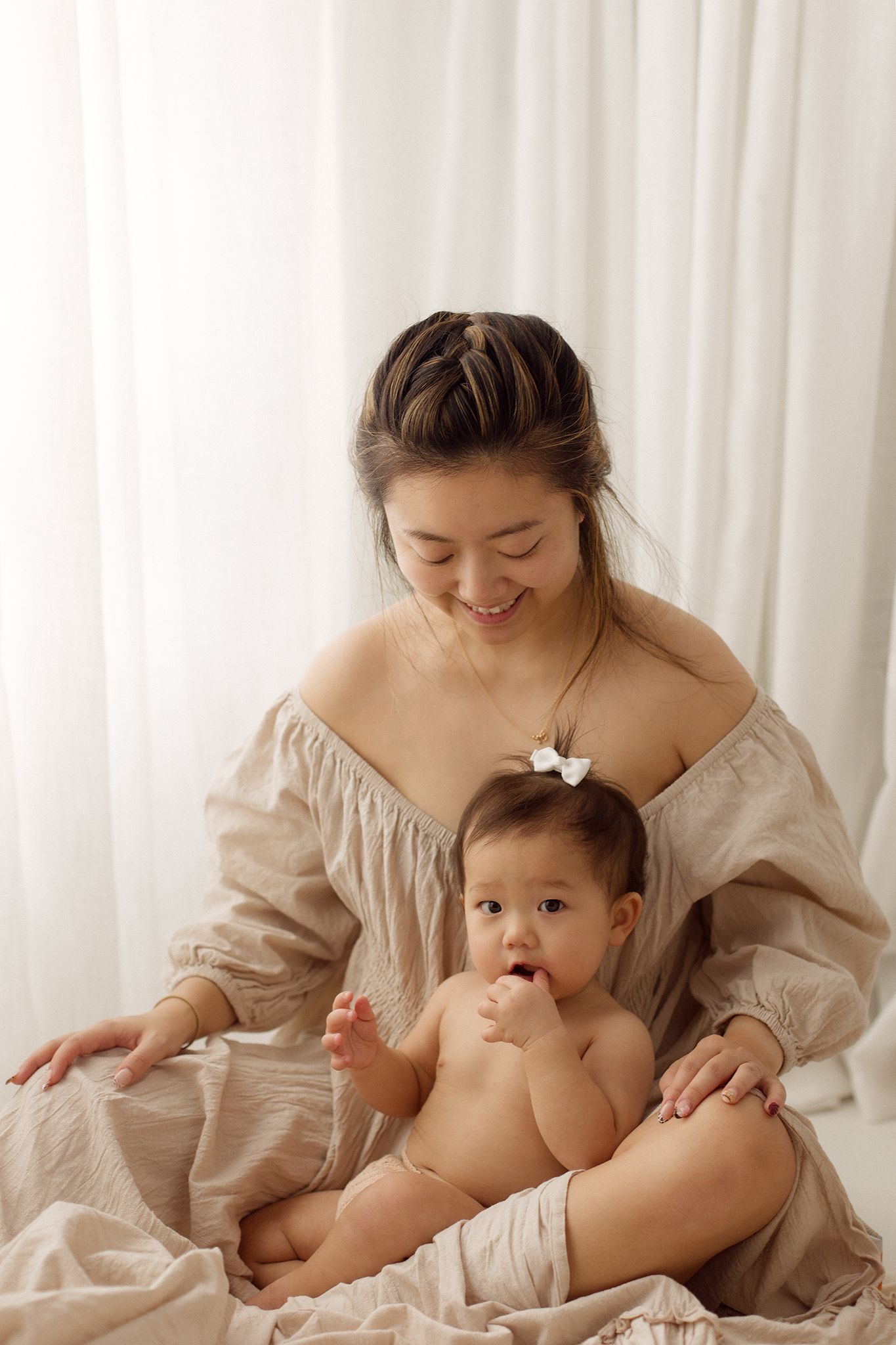 Smiling mother in a cream off-shoulder top sits with her baby, who wears a small bow, against soft white curtains in a bright studio.