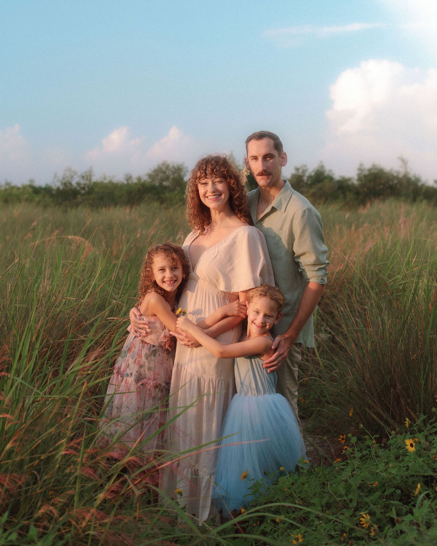 A smiling family of four poses at sunset in a tall green grass field, with two young girls in dresses hugging their parents.