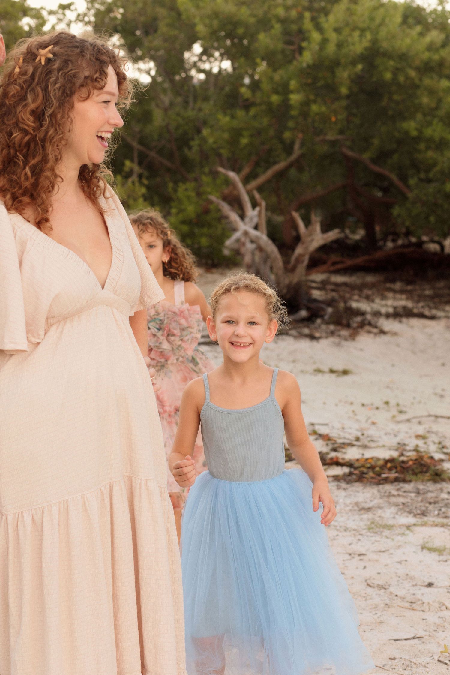 A smiling girl in a blue tulle dress stands on a sandy beach beside a laughing woman in a cream dress, with mangrove trees behind them.