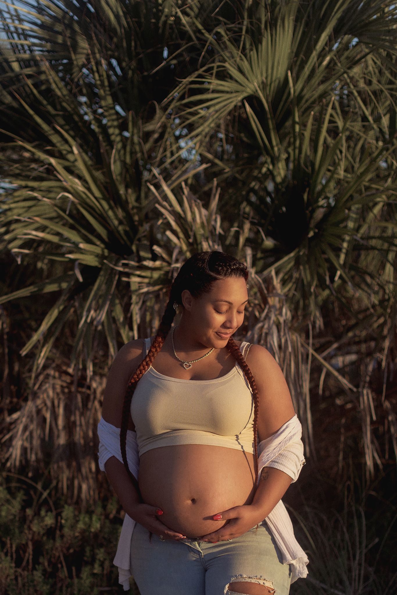 Pregnant woman in white crop top and jeans cradling baby bump outdoors with tropical palm trees in warm golden sunlight.