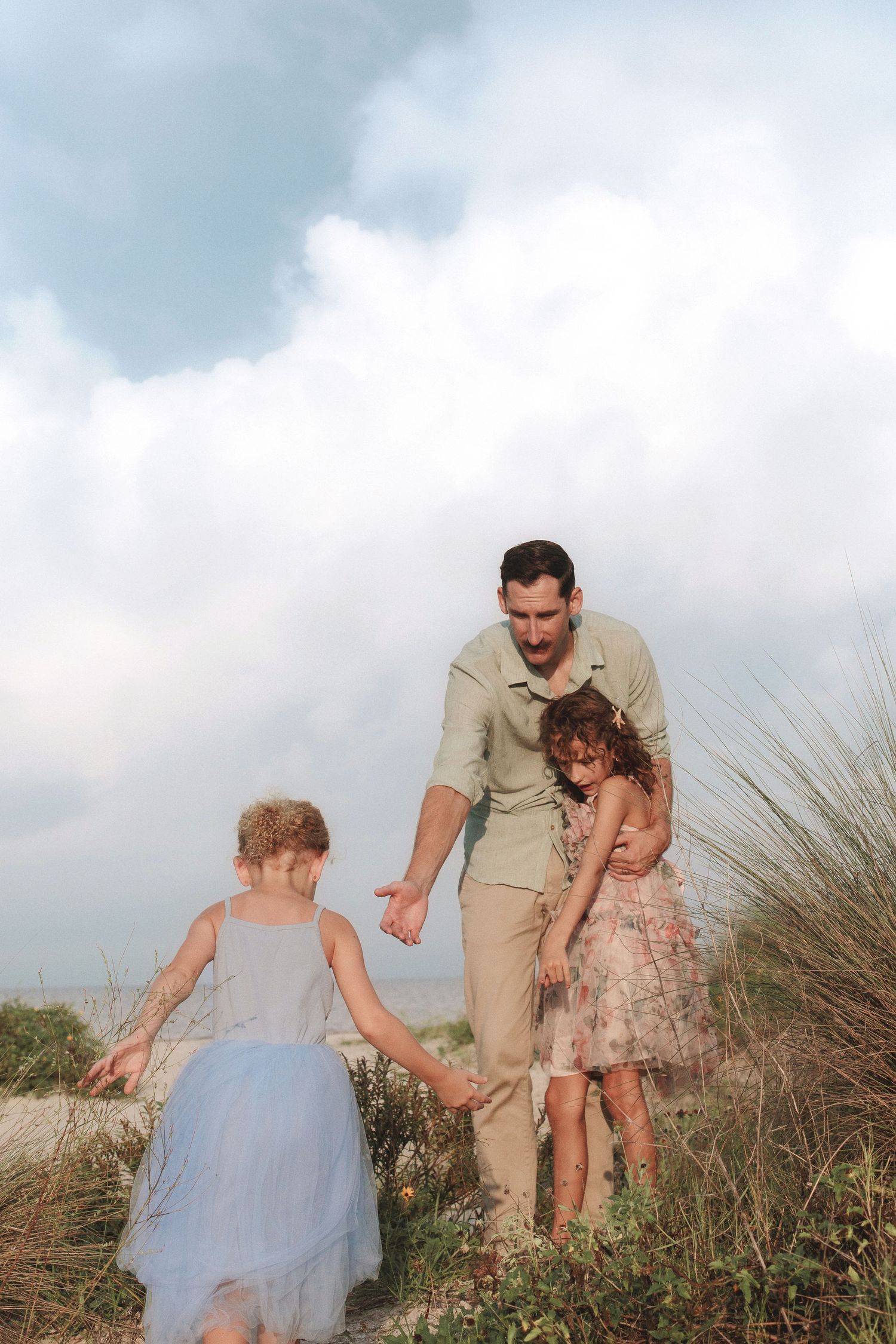 Father in khaki outfit hugs a young girl while another girl in a blue tulle skirt reaches out on a grassy beach dune under a cloudy sky.