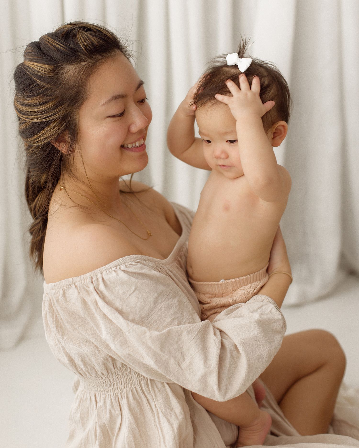 Smiling woman in a cream off-the-shoulder blouse holds a baby wearing a white hair bow against a soft white curtain backdrop.