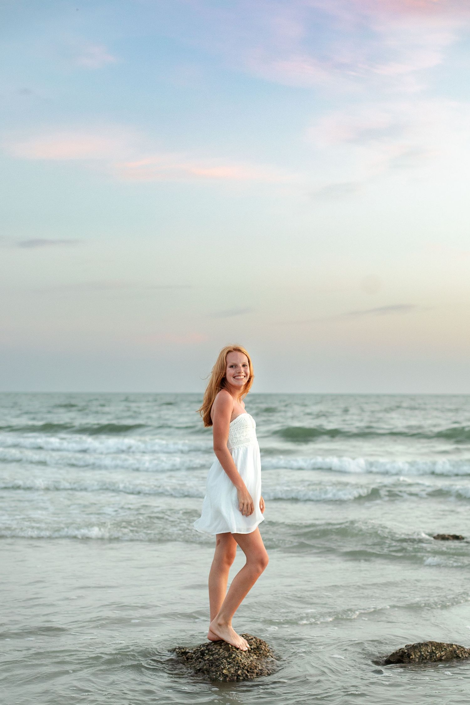 Young woman in white dress standing on rocks at beach during sunset with ocean waves in background.