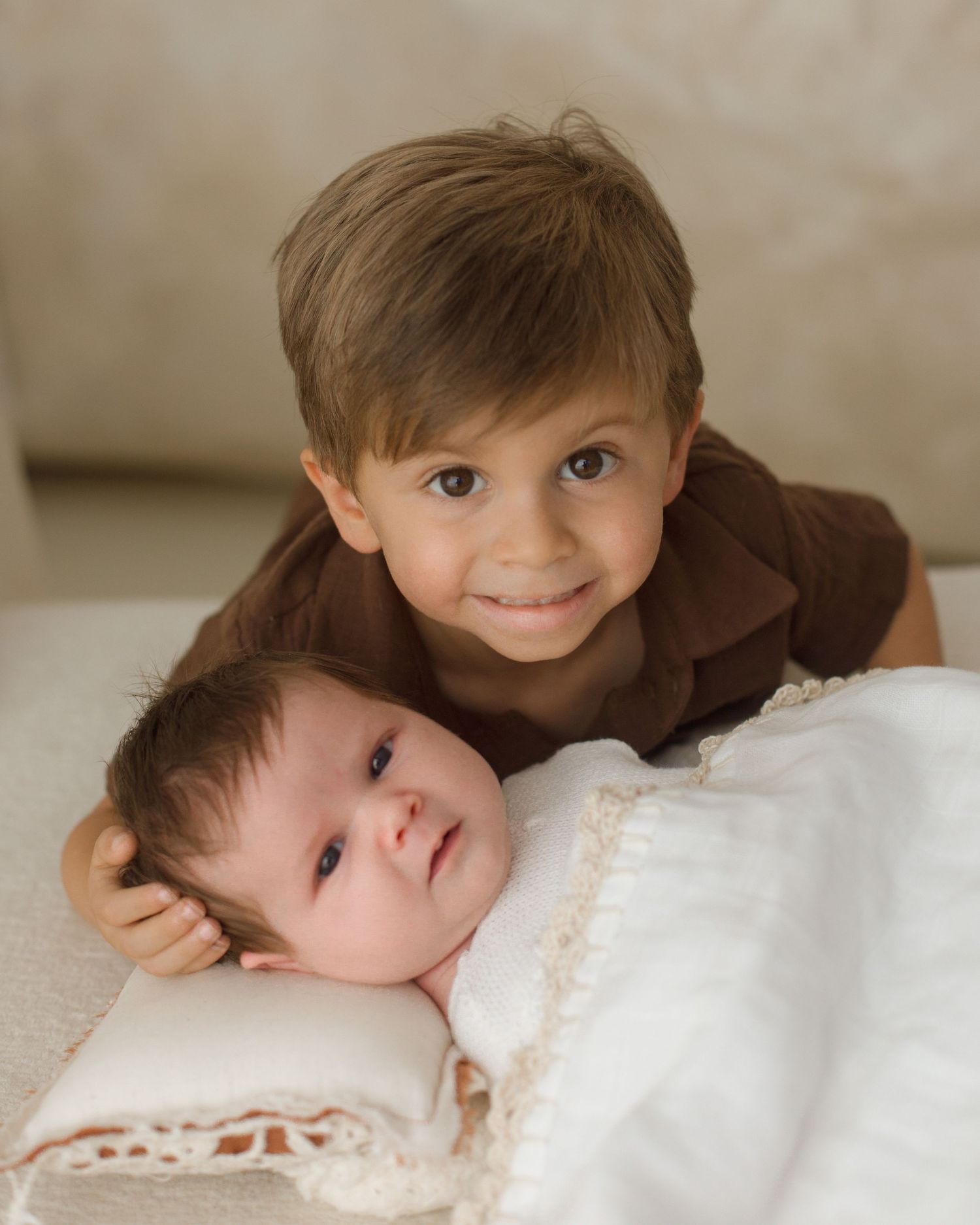 A smiling young boy gently rests his arm around a newborn baby wrapped in a white lace blanket.