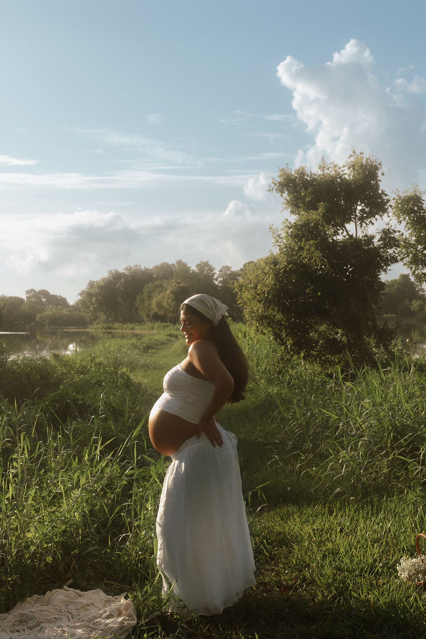 Pregnant woman in white dress and headband standing in lush green meadow near water under a bright cloudy sky.