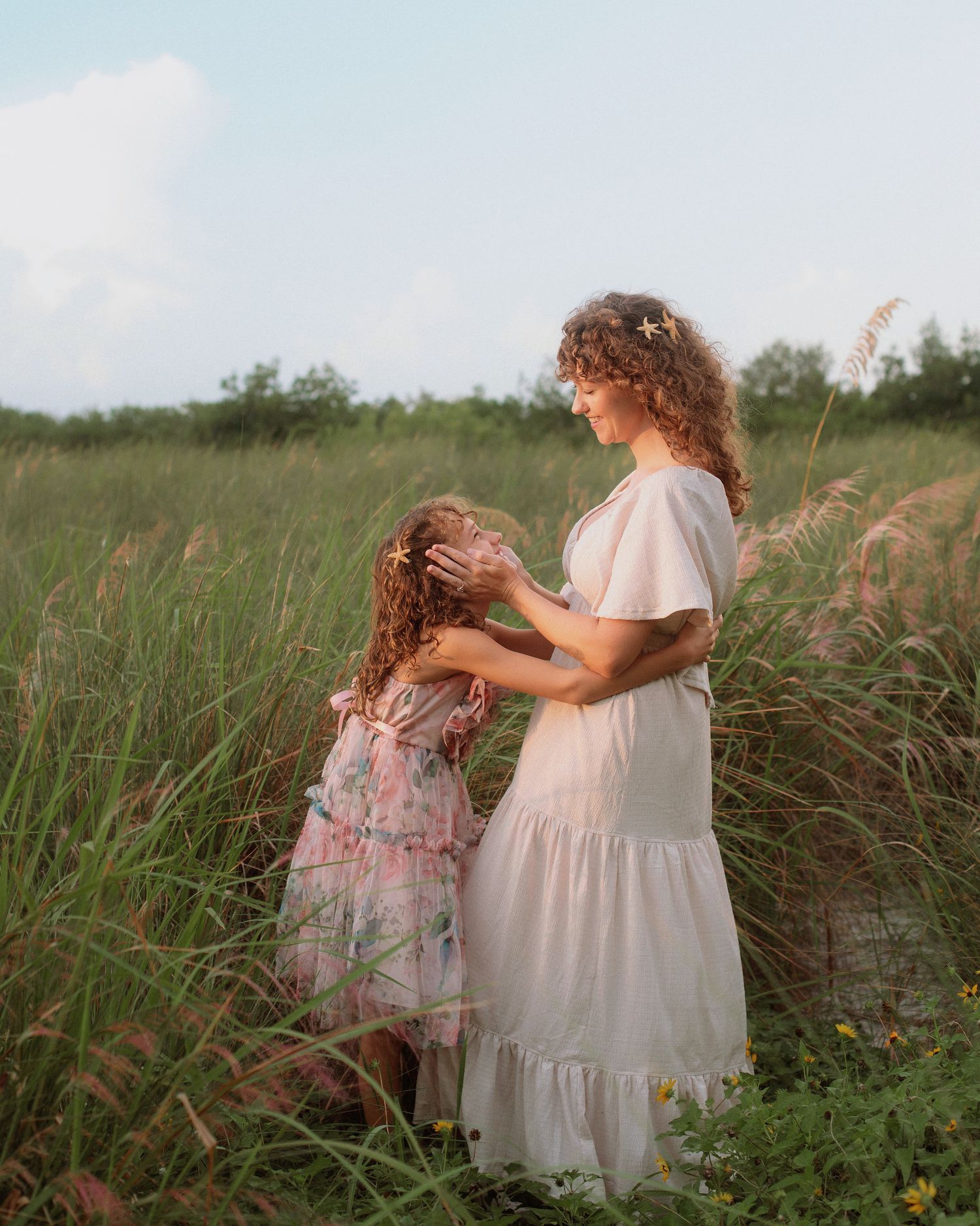 A woman in a cream dress gently touches the head of a young girl in a floral dress amid tall green grass at golden hour.