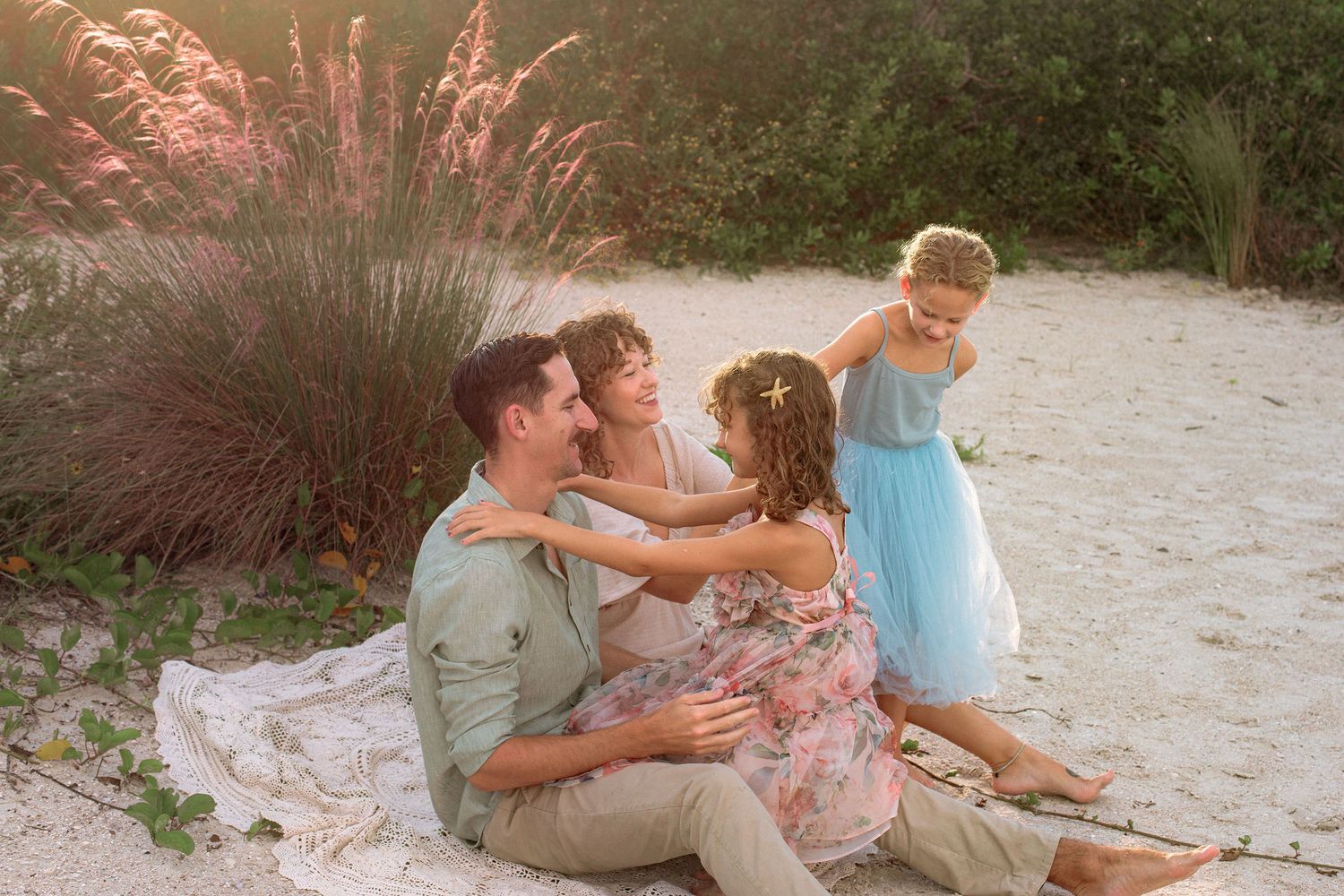 Family of four sitting together on a white blanket on a sandy beach at sunset, with pink pampas grass and greenery in the background.