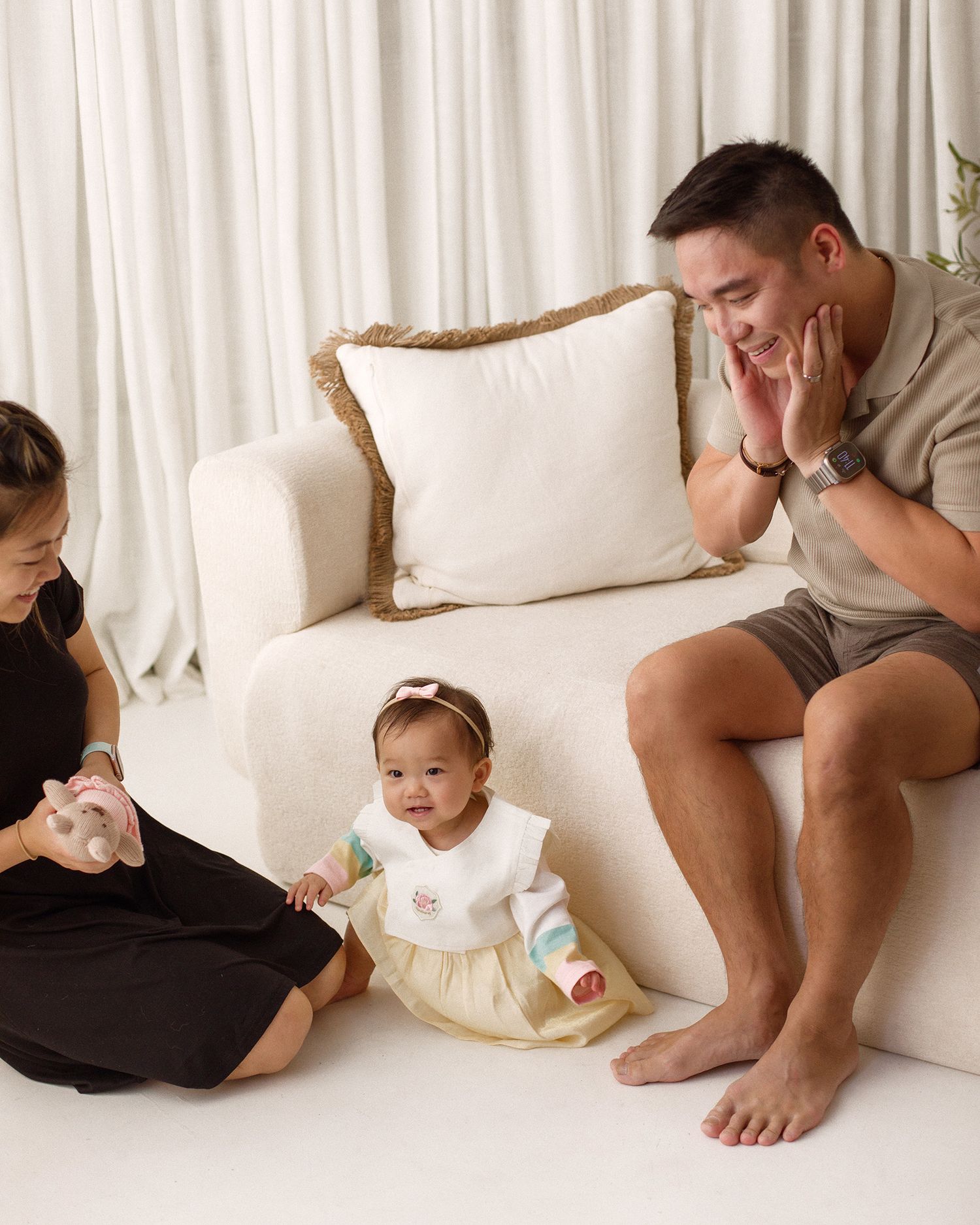 A smiling father sits on a white sofa while a baby in a traditional outfit sits on the floor beside a mother in a black dress.