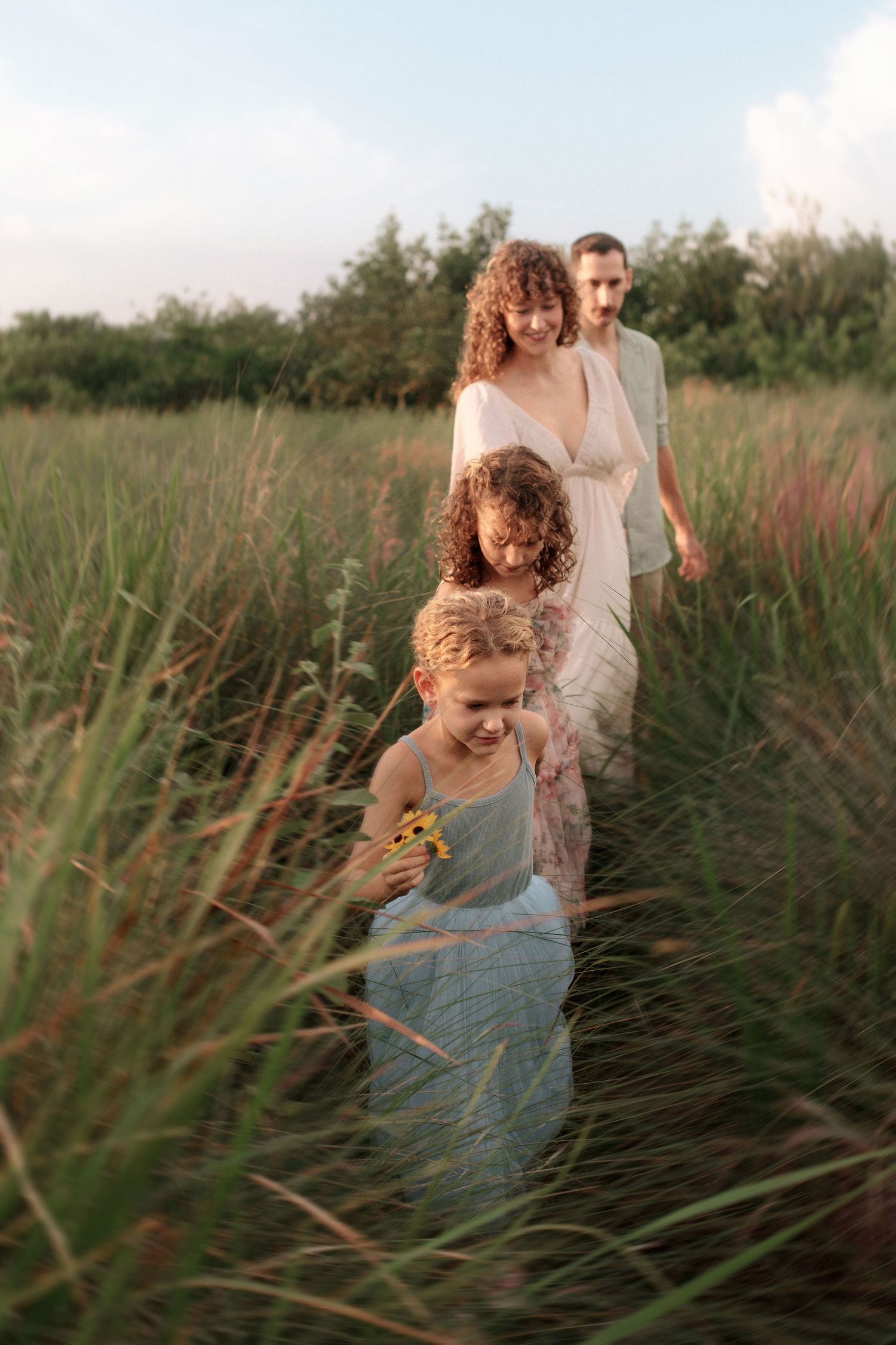 A family of four walks single file through tall golden grass in a sunlit field, a young girl holding wildflowers leading the way.